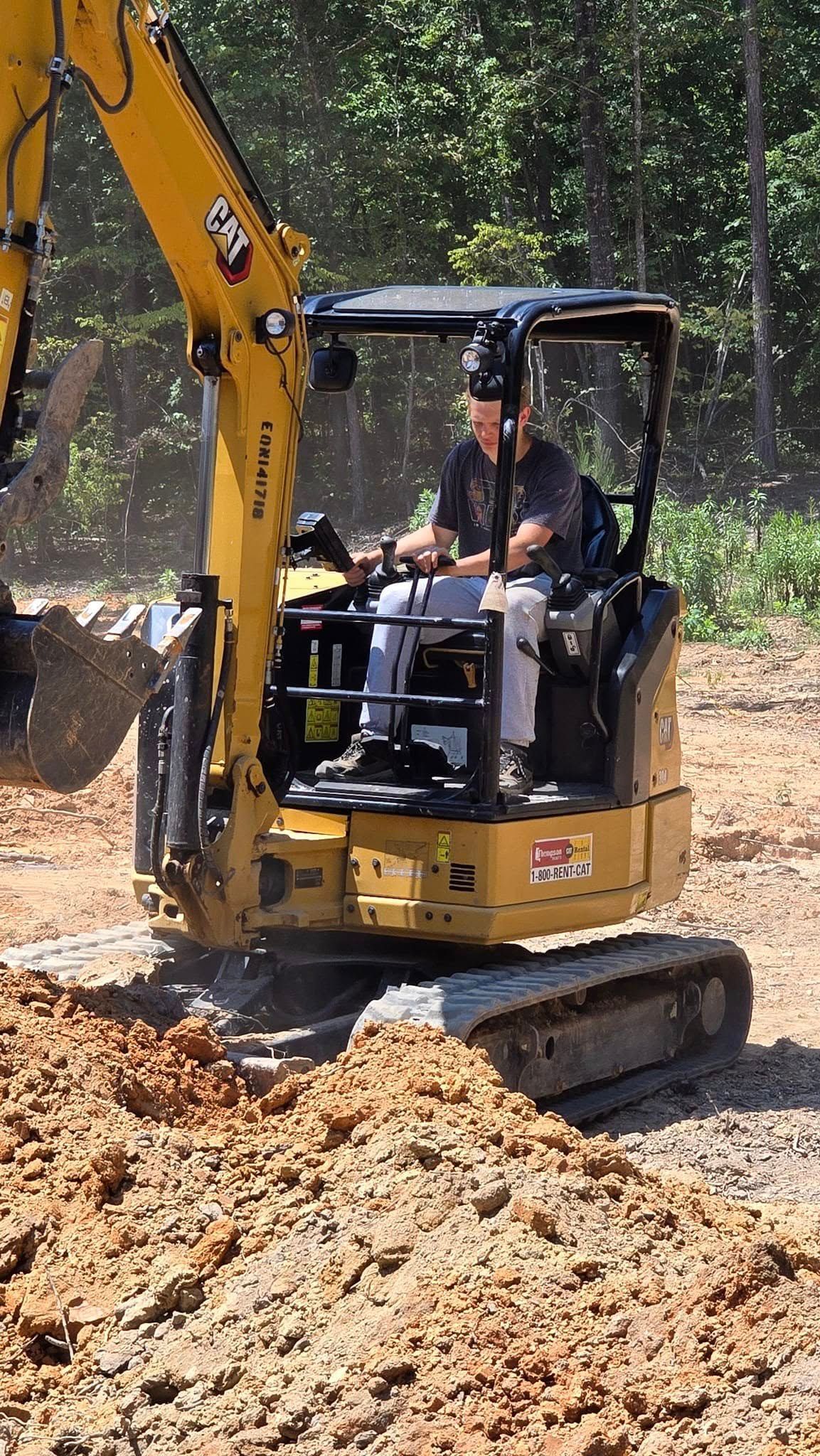 Yellow excavator digging dirt; person operating it. Outdoors on a sunny day.