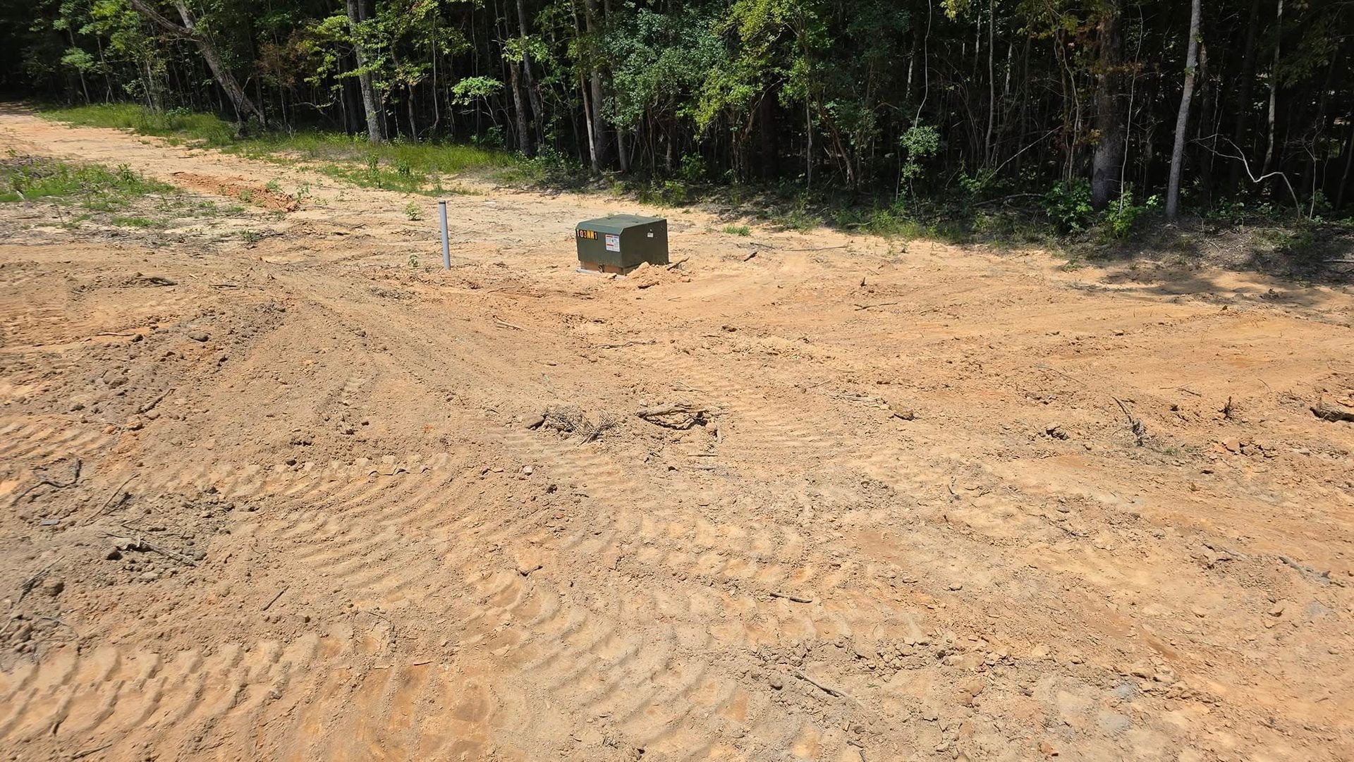 Dirt road with tire tracks, a dark box sits in the center, trees in the background.