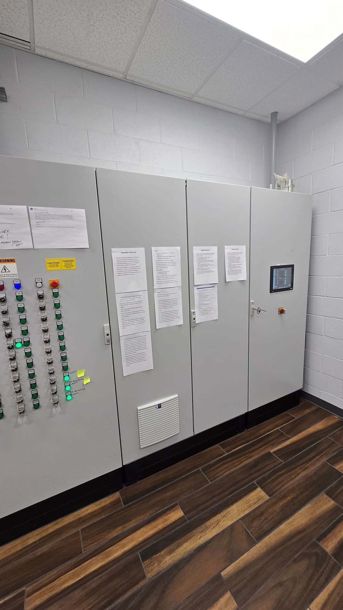 Control panel with various buttons, lights, and documents mounted on a wall. Brown floor, white ceiling, and gray panels.