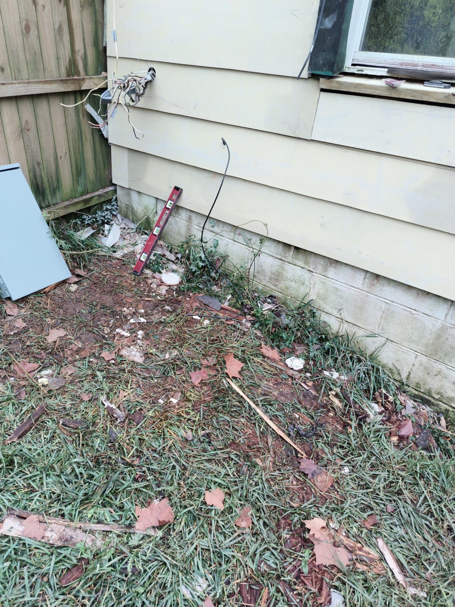Damaged house exterior with exposed wires near a window and dirty ground covered with leaves and debris.