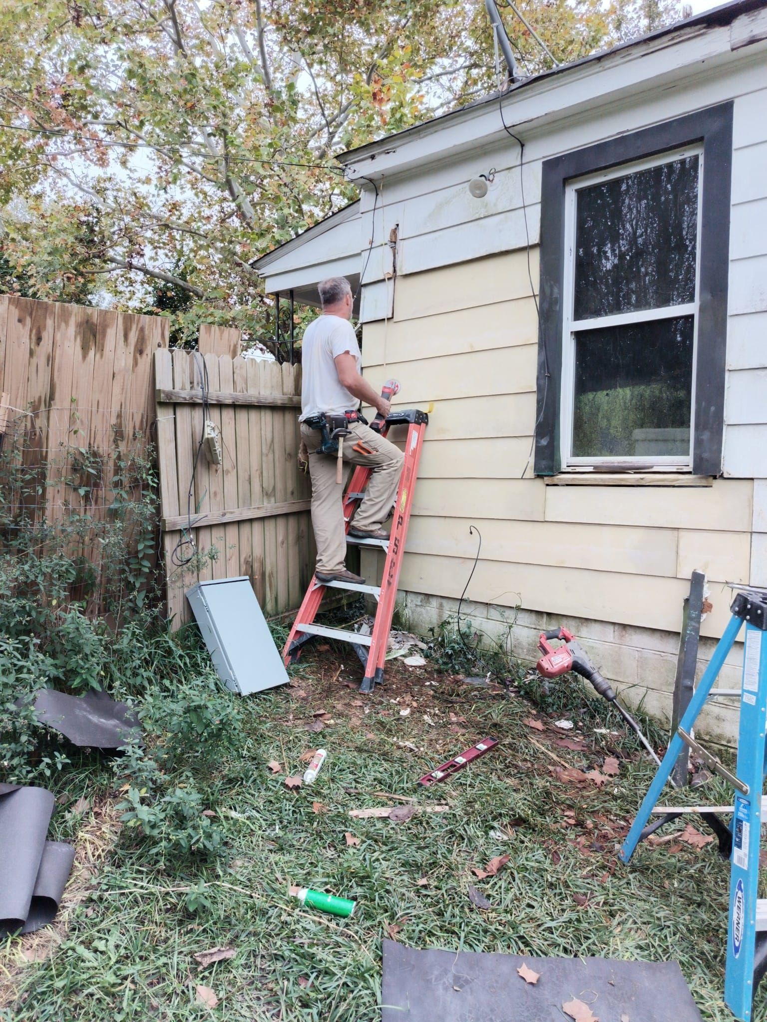 Man on a ladder repairing siding of a house next to a wooden fence.