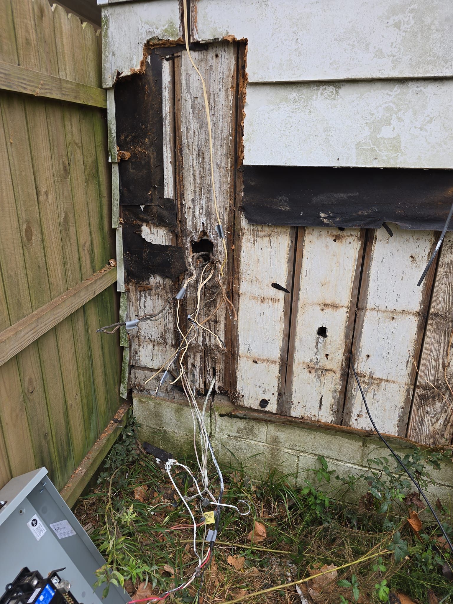 Damaged exterior wall with exposed wiring and missing siding next to a wooden fence.