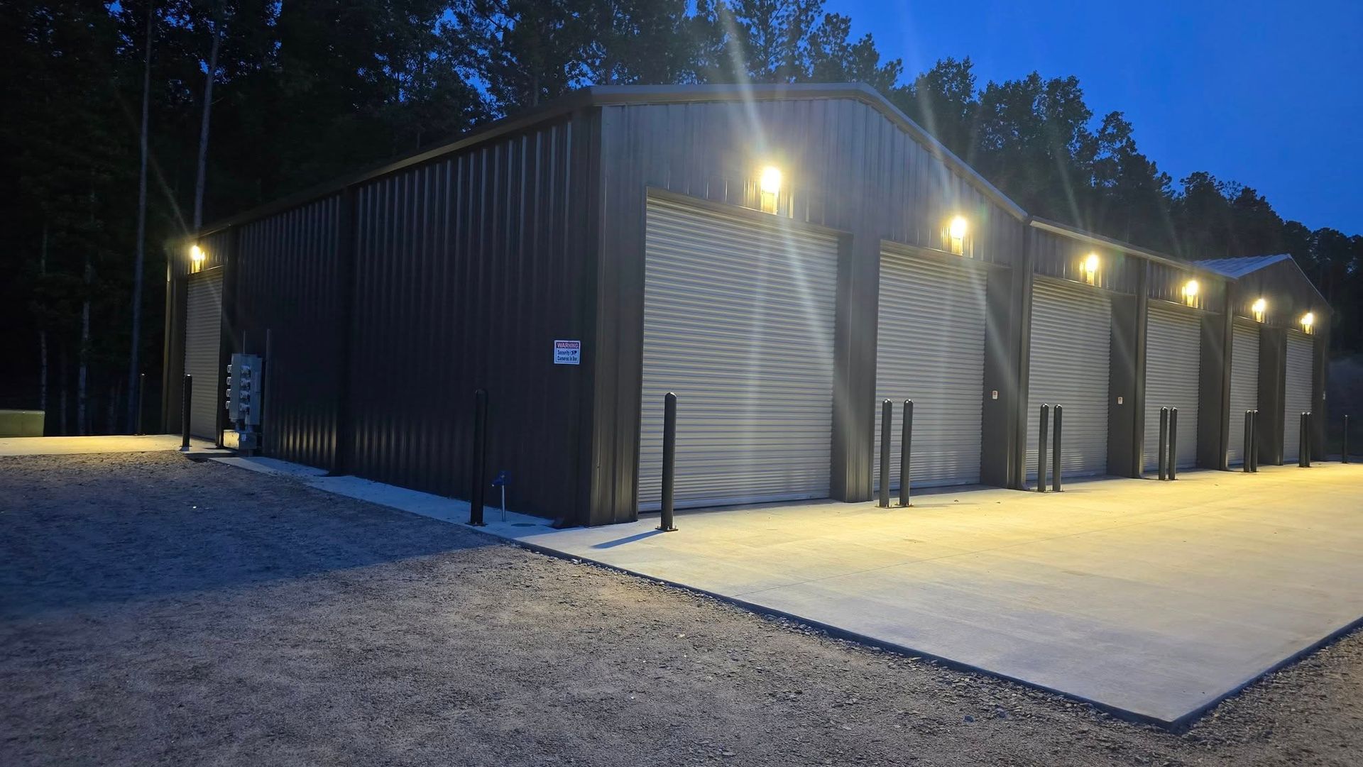 Dark brown metal building with multiple bays, lit by exterior lights, gravel and concrete.