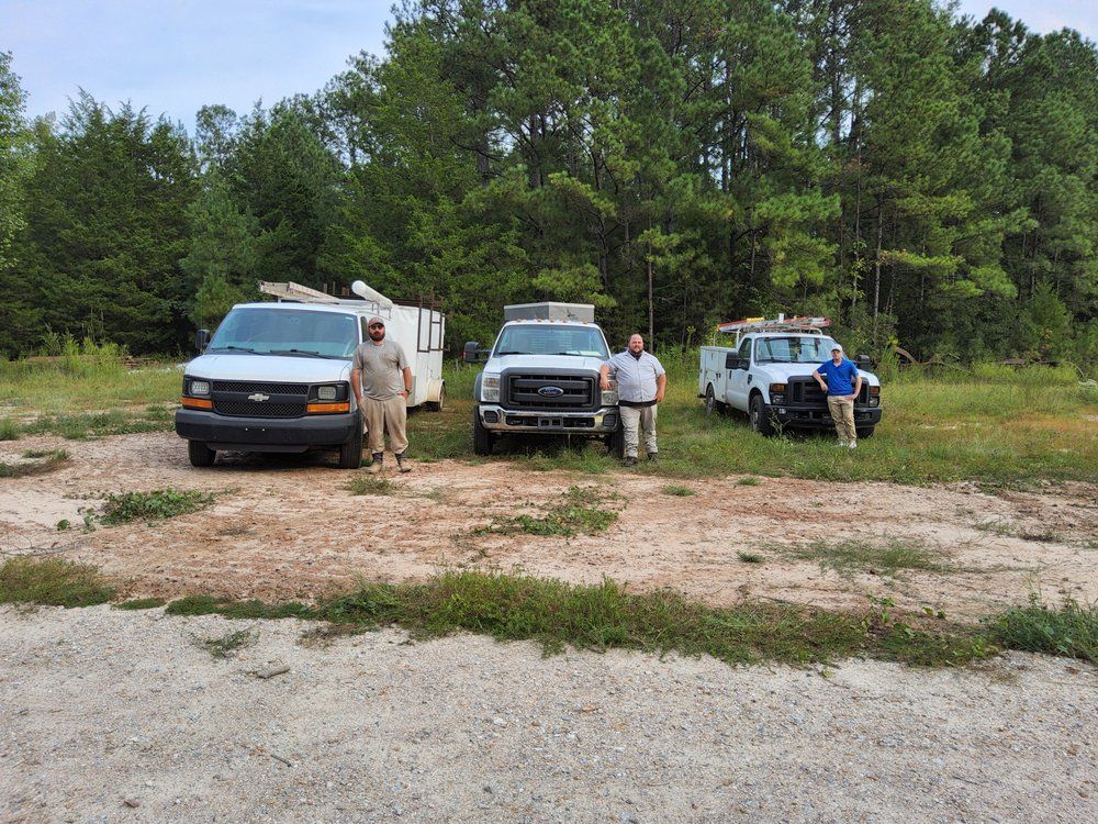 Three people stand near work trucks in a clearing. Vehicles are white and gray. Trees are in the background.