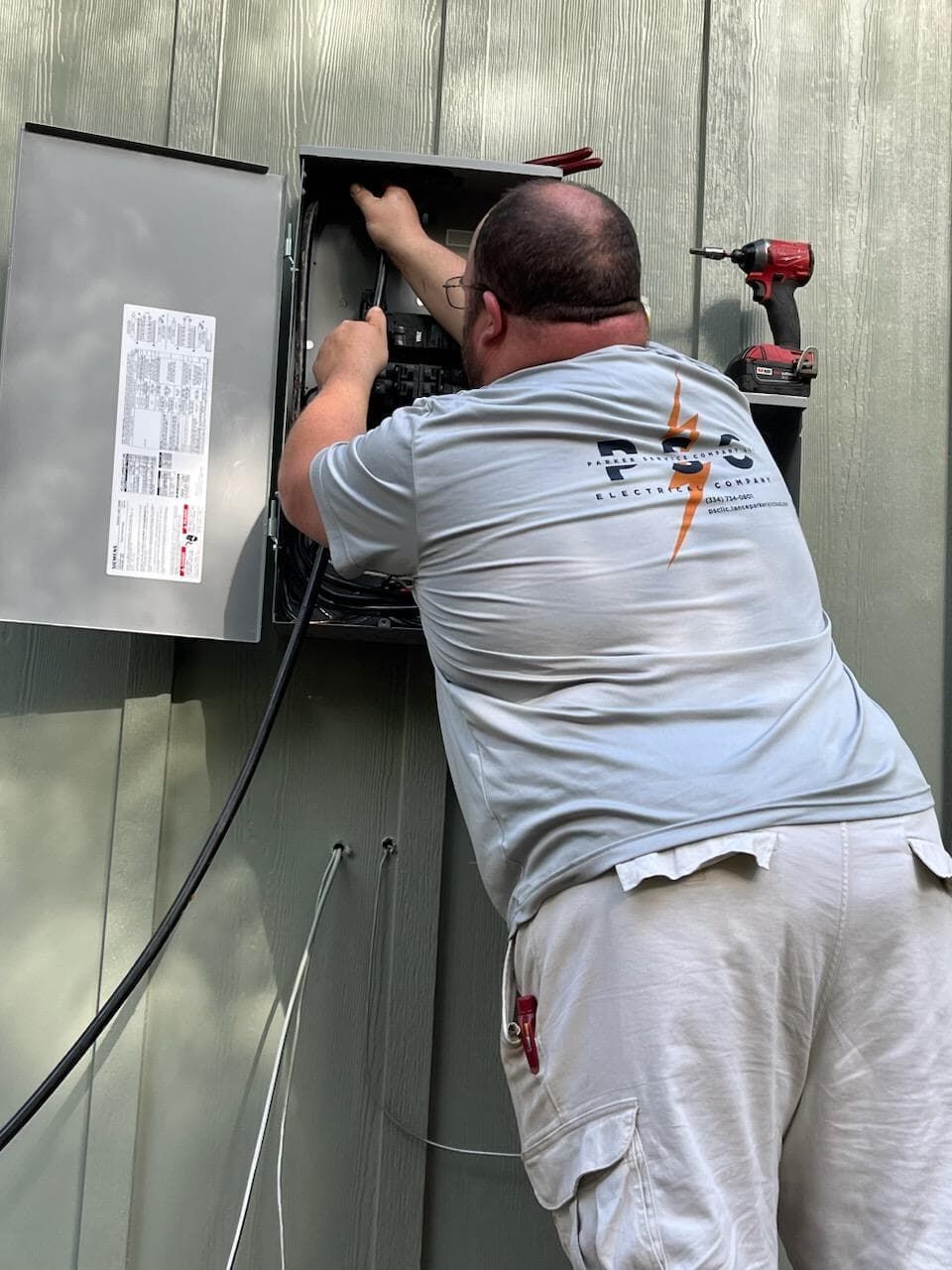 Electrician working inside an electrical box mounted on a gray siding wall, using tools.
