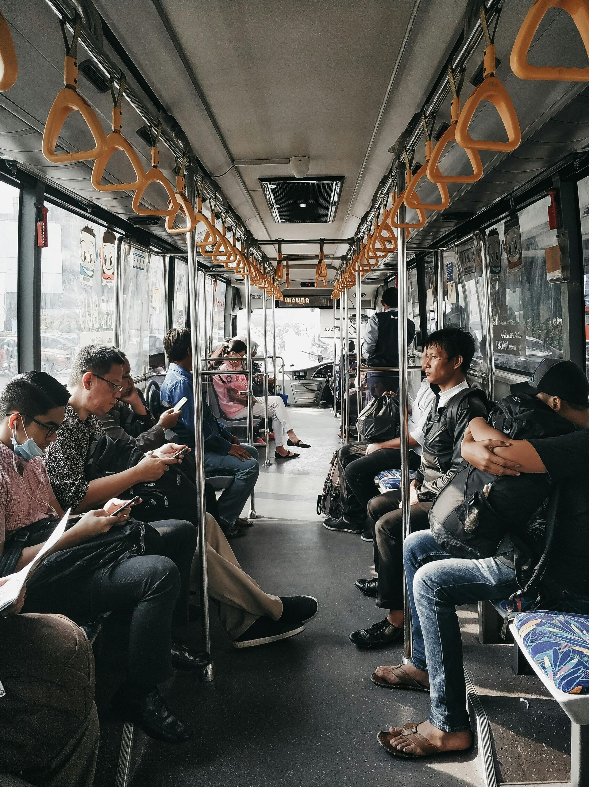 Interior of a bus, several people seated and standing. Overhead handrails are yellow. Sunlight streams in from windows.