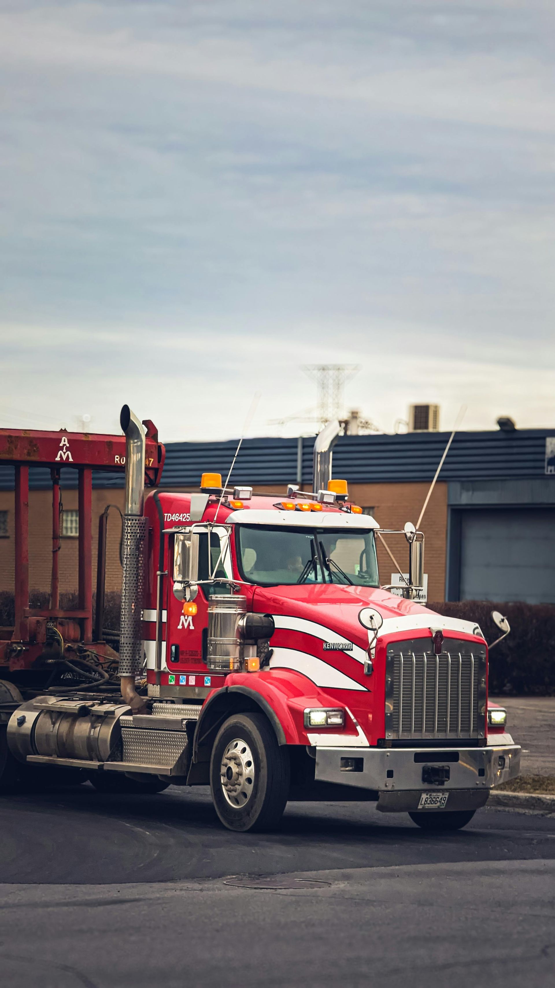 Red semi-truck parked on a gray road. The truck has chrome details and a white stripe design. Background is a building and cloudy sky.