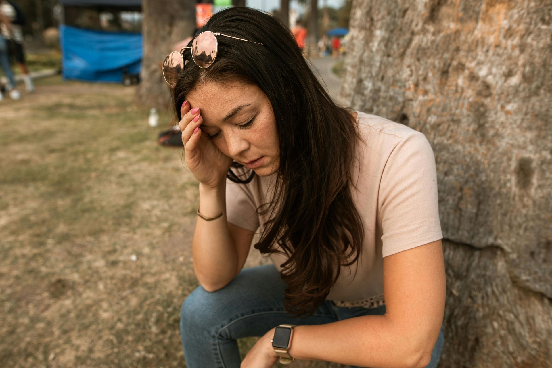 Woman with head in hand, sitting near a tree, looking distressed outdoors.