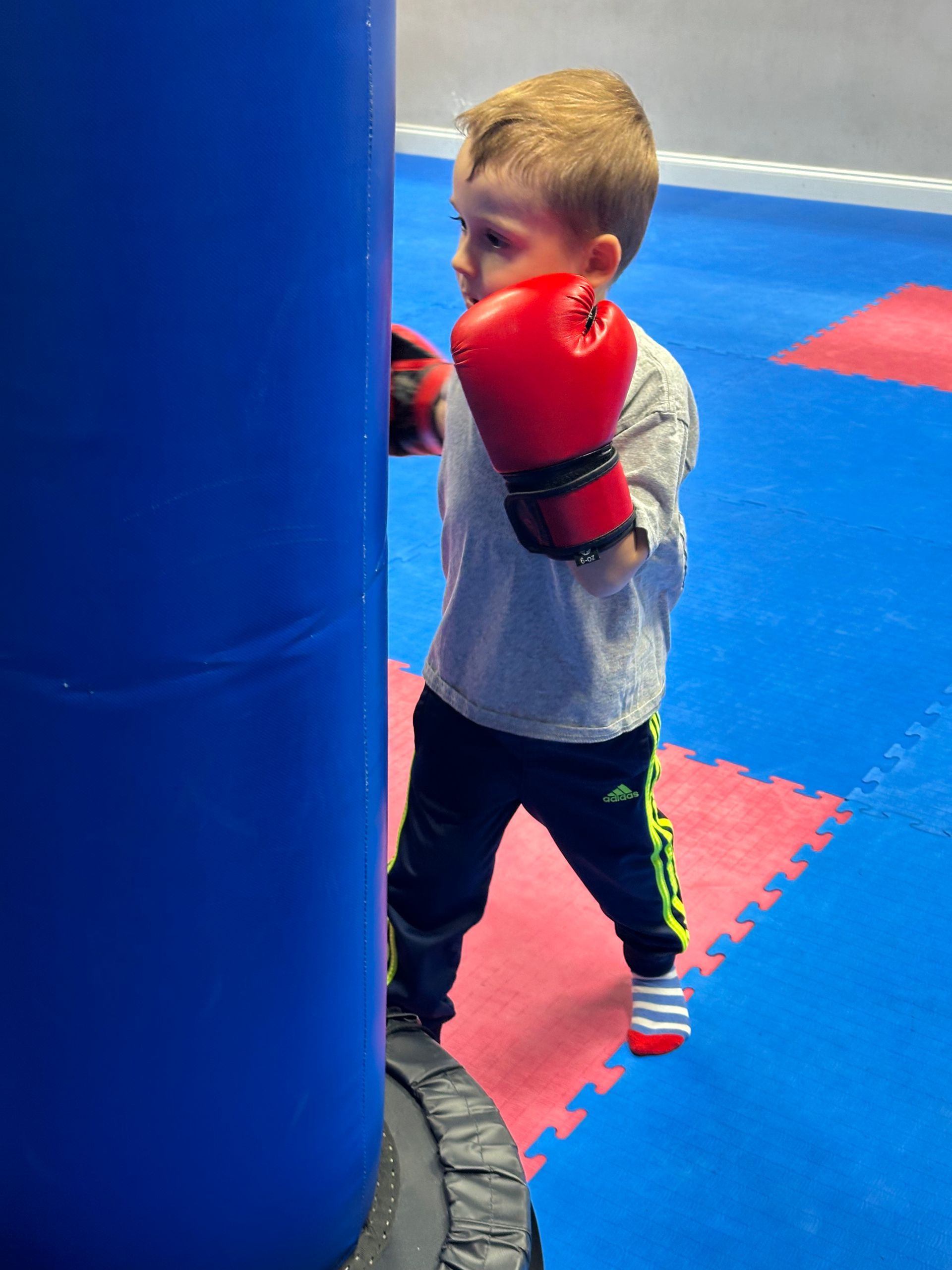 Child wearing red boxing gloves practices punches beside a blue padded pillar in a gym.