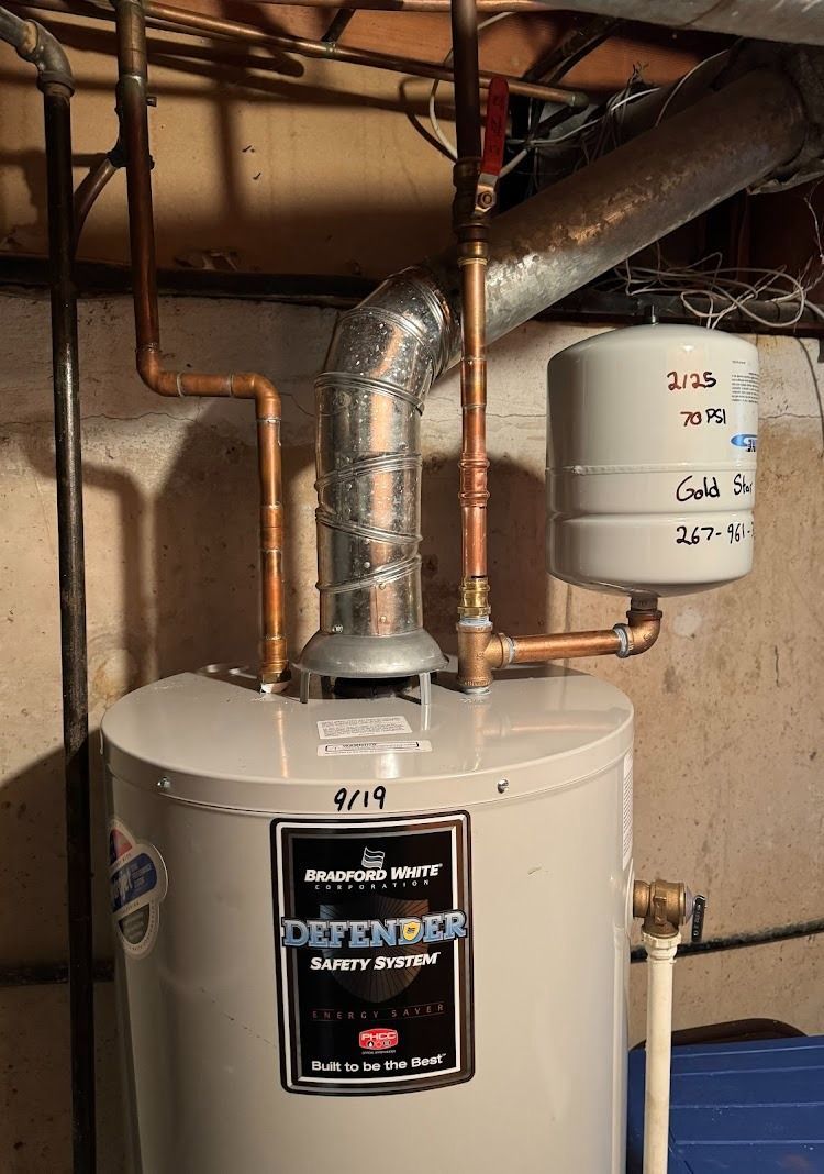 A water heater with copper pipes and a cylindrical expansion tank in a basement setting.