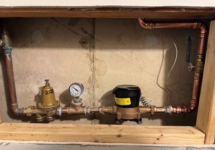 Copper pipes and water meter in a framed wall cavity. A pressure regulator and gauge are visible.