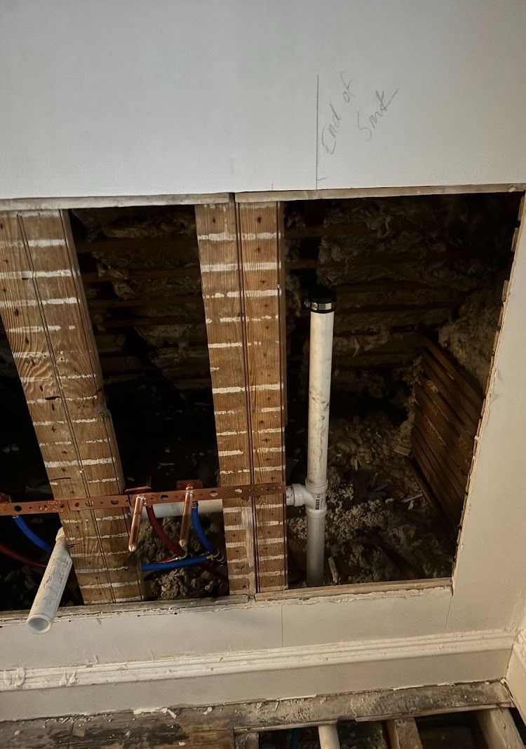 Hole in a ceiling revealing plumbing and wooden beams. White pipes, blue/red water lines, and insulation are visible.