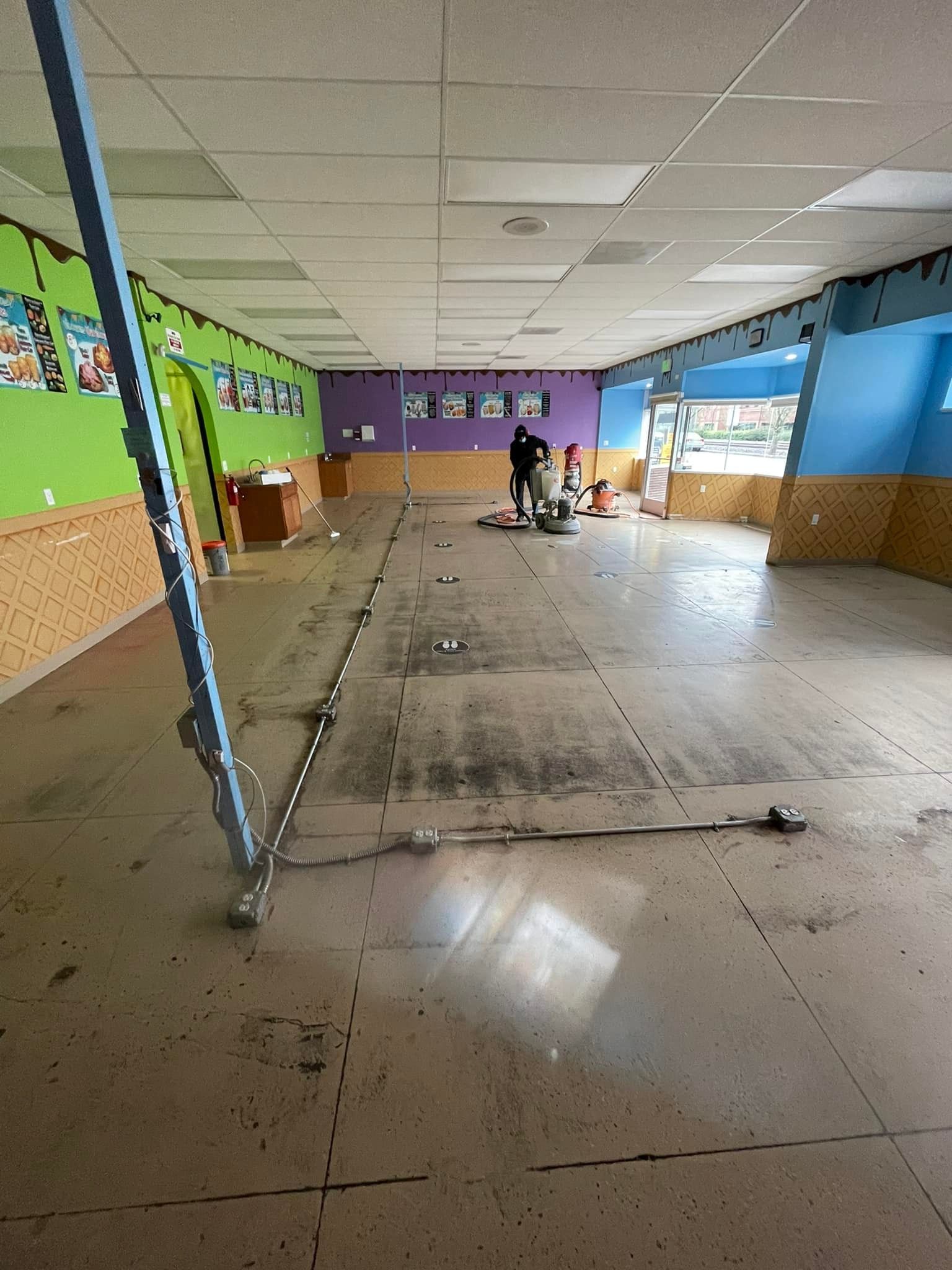 Elite Surface Coatings - Person sanding a light brown floor in an empty room with colorful walls, a blue support beam.
