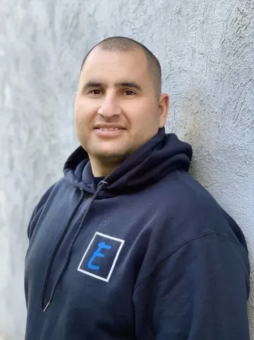 A man in a blue hoodie is leaning against a wall and smiling.
