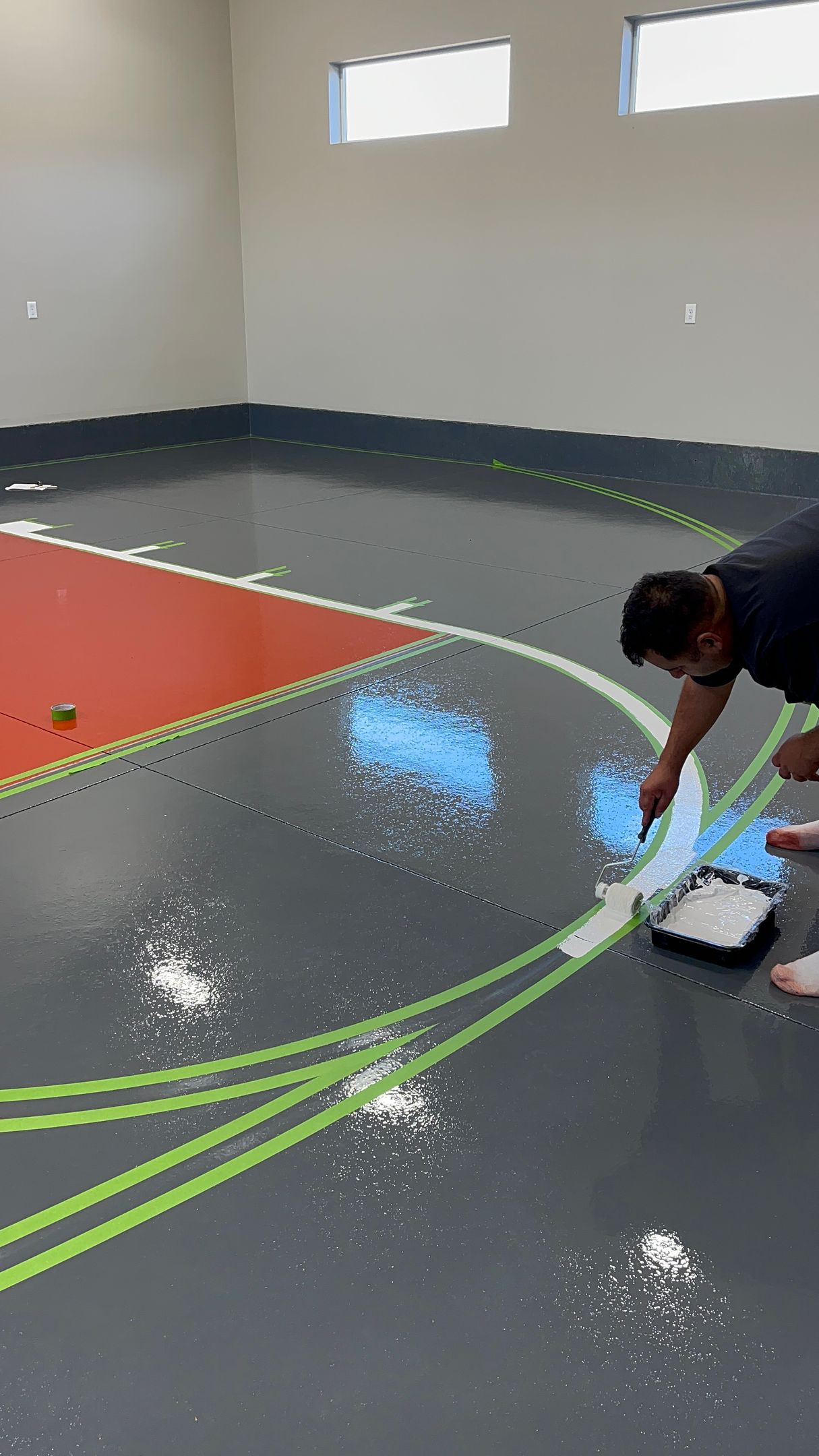Elite Surface Coatings -Man painting blue area inside a basketball court's key. The floor is gray with green lines. Red paint is visible.