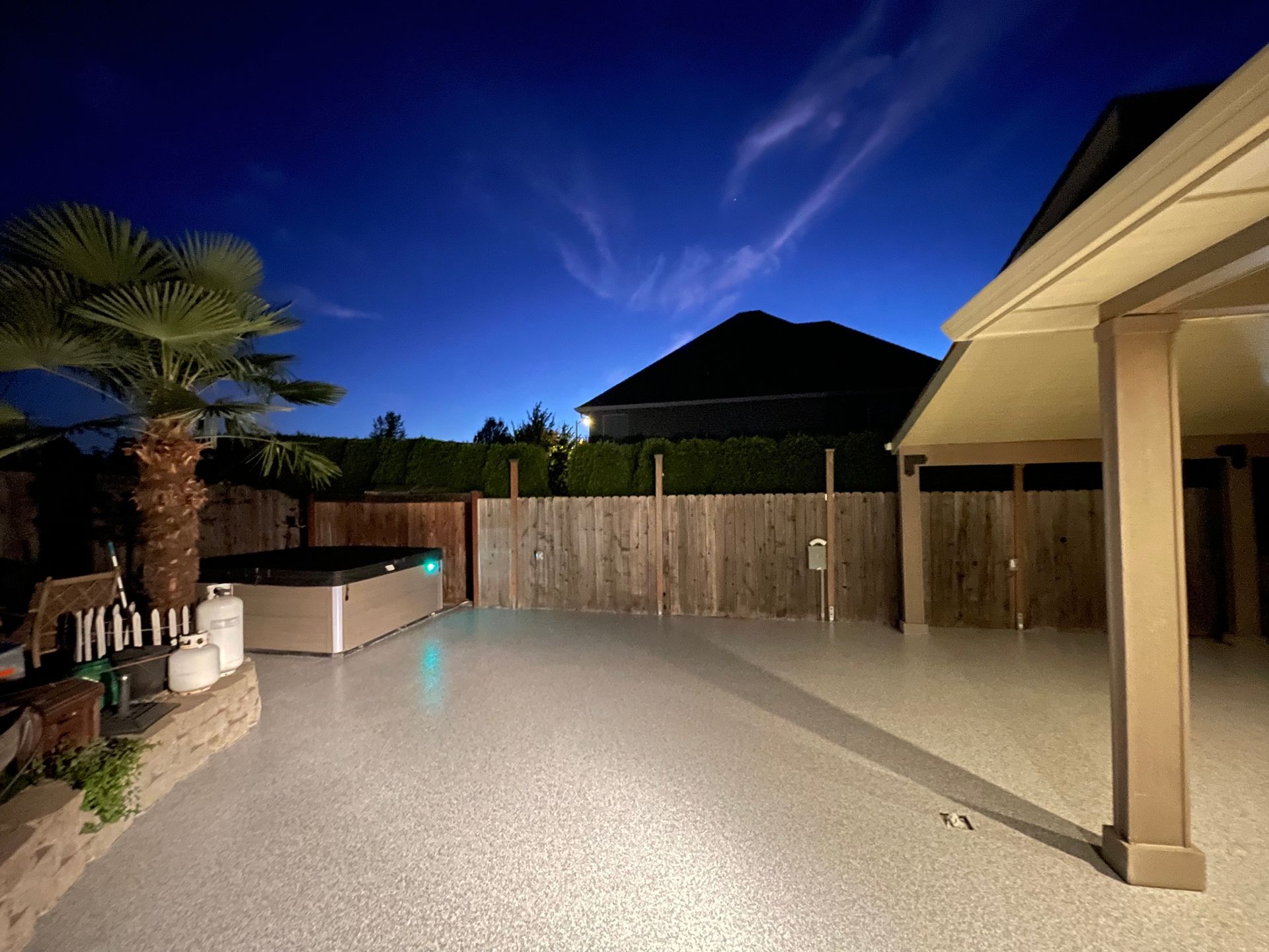 Elite Surface Coatings - Backyard at dusk: hot tub, palm tree, covered patio, wooden fence, blue sky.
