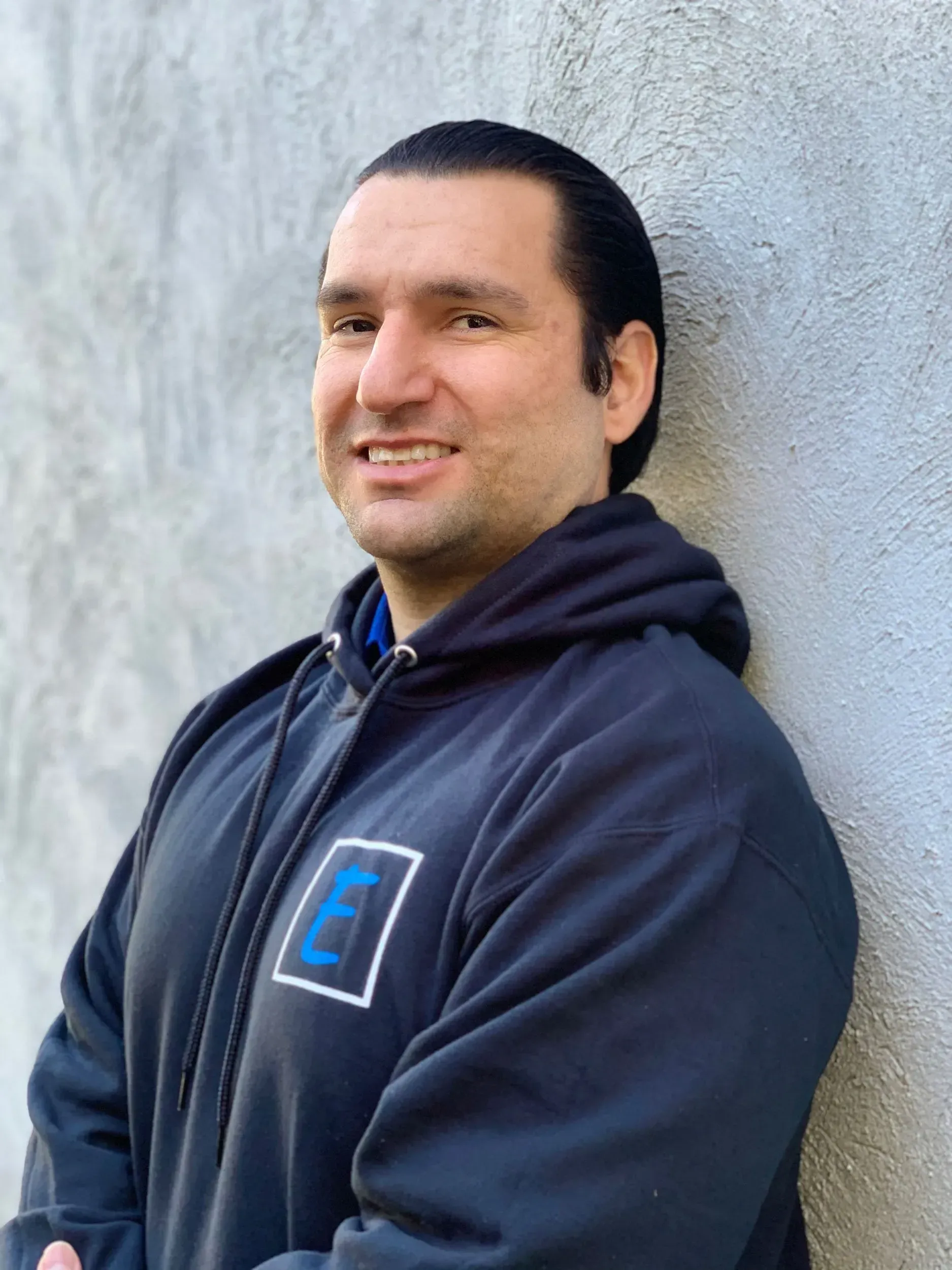 A man in a black hoodie is leaning against a wall and smiling.