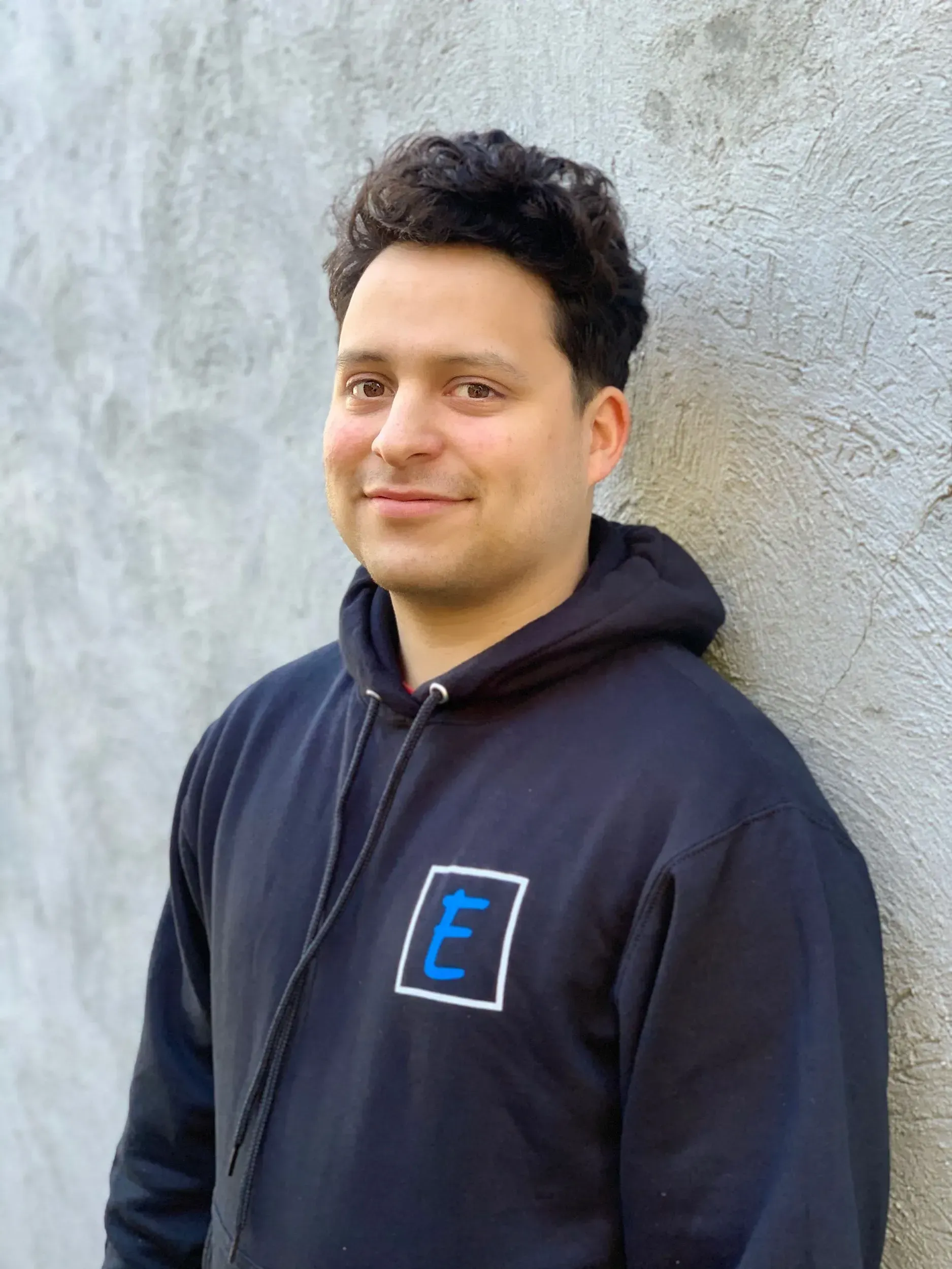 A man wearing a black hoodie with the letter e on it is standing in front of a wall.
