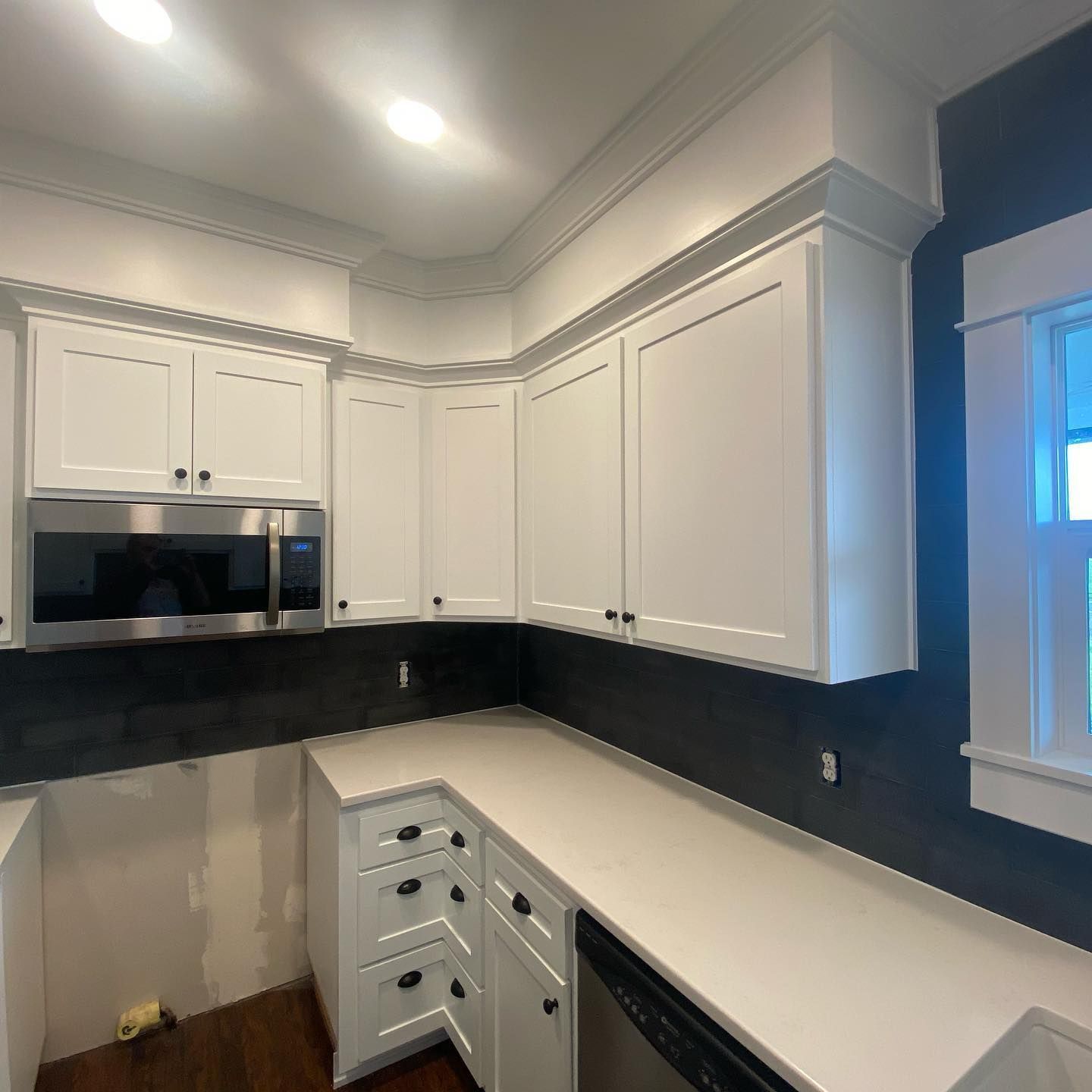 Elite Surface Coatings – White kitchen cabinets and countertop, dark backsplash, stainless steel microwave, and window.