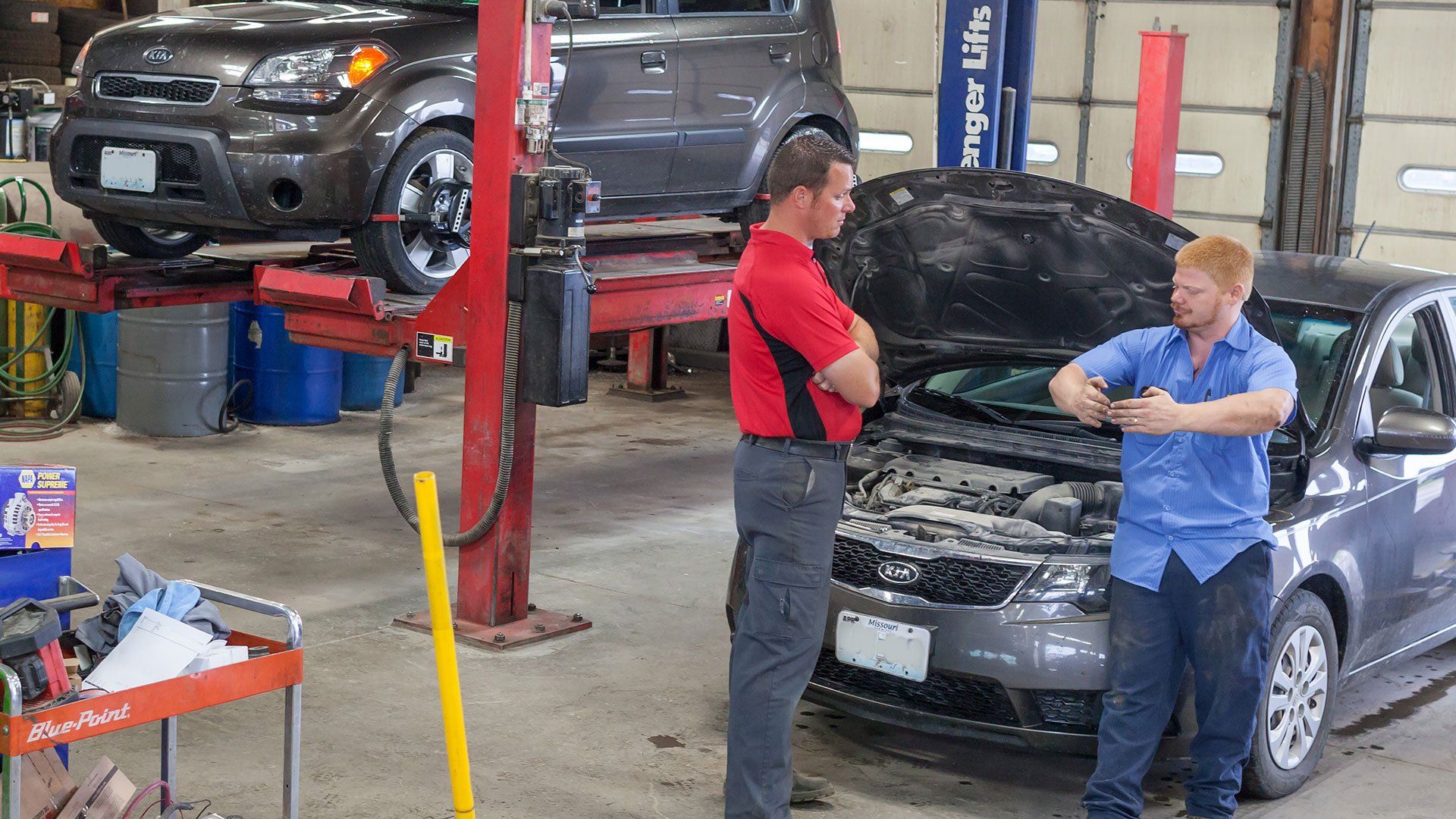 Two men talk in an auto shop beside a gray car with its hood open; another car is on a lift in the background.