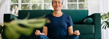 A woman in meditation, eyes closed, sits cross-legged in front of a teal sofa.