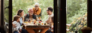 Family gathered around a table outdoors; sharing a meal. Older couple, adults, child. Green trees in the background.