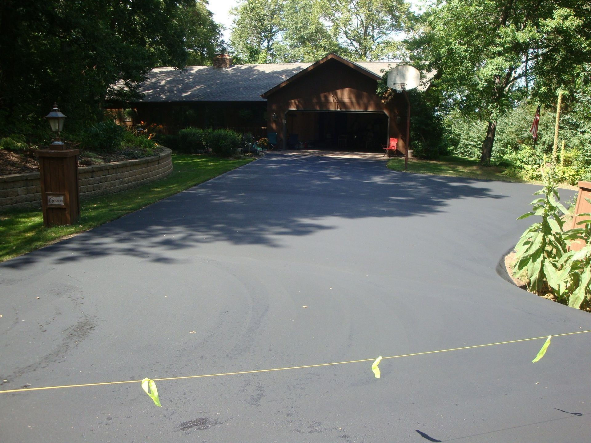 Black asphalt driveway leading to a brown house with a garage and basketball hoop, surrounded by trees.