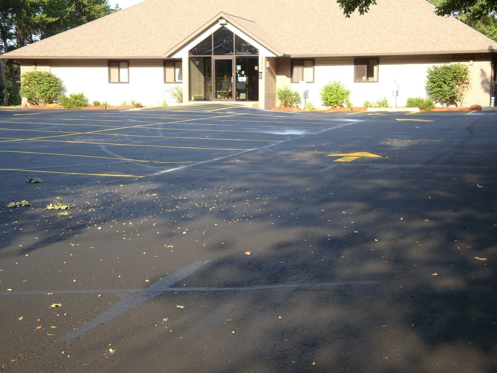 Building with a parking lot. Brown roof, glass entrance, black asphalt with leaf debris.