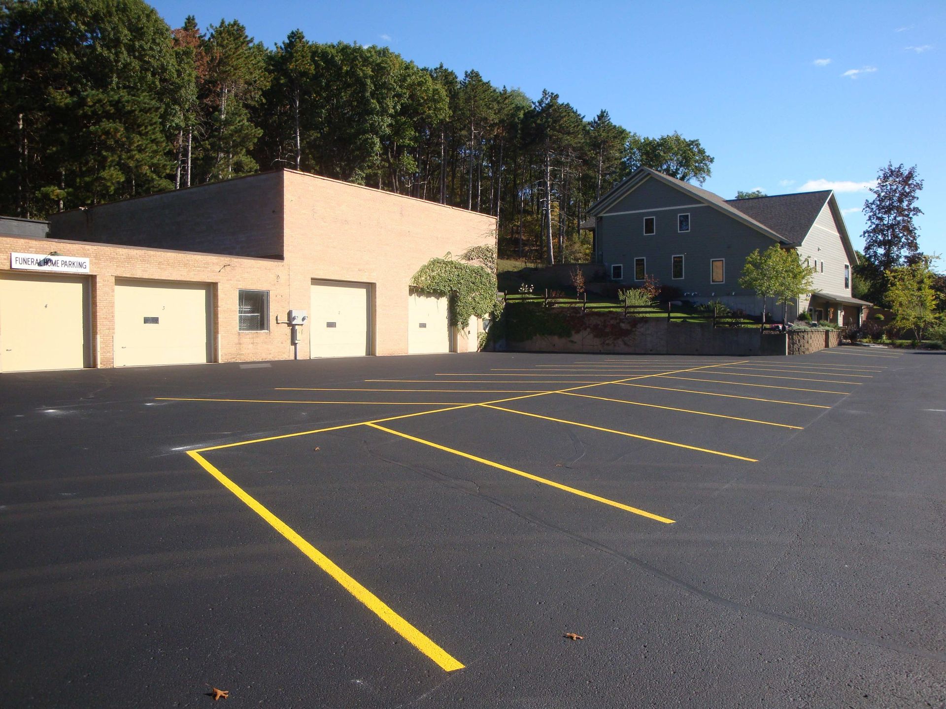 Parking lot with yellow lines, beige building with garage doors, and a gray house, trees in background.