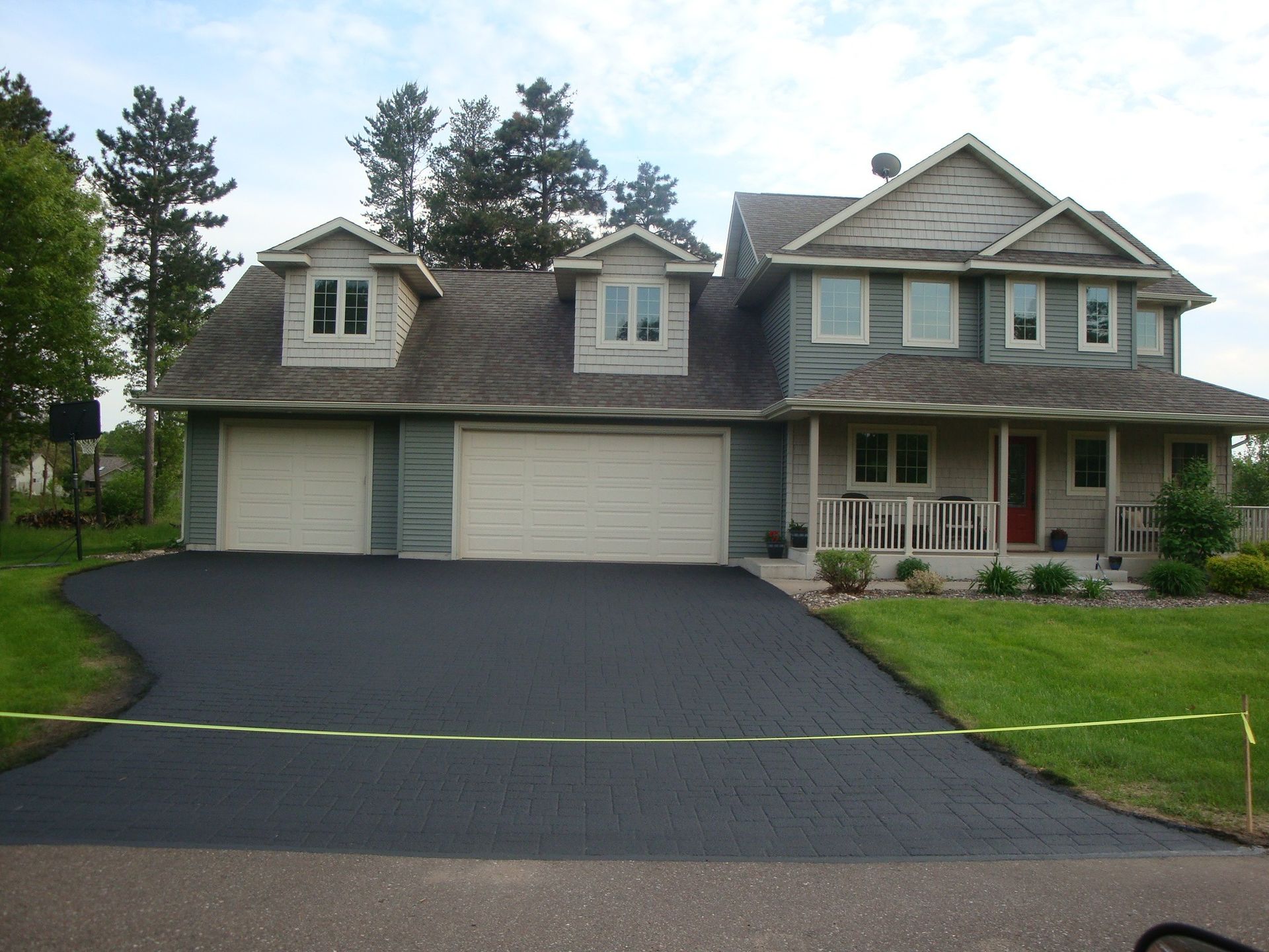 Two-story house with black asphalt driveway. Green grass, beige garage doors, and dormers.
