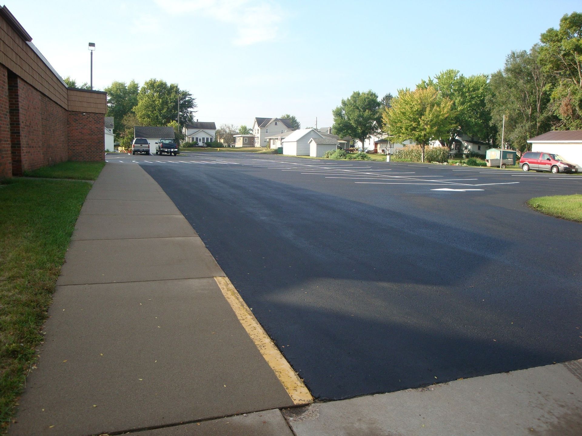 View of newly paved parking lot with sidewalk, brick building and houses in the distance.