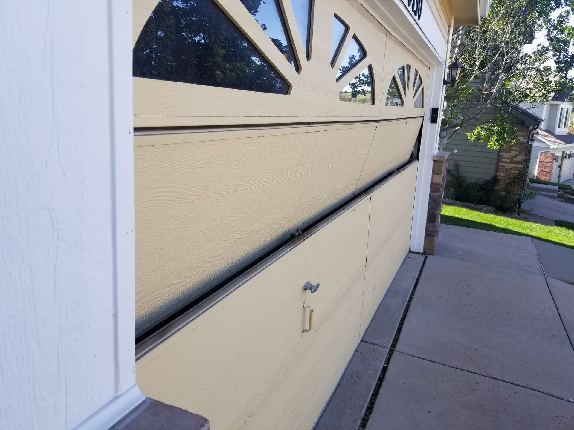 Garage door, partially open, angled view. Beige door with dark trim, sidewalk, and green grass.