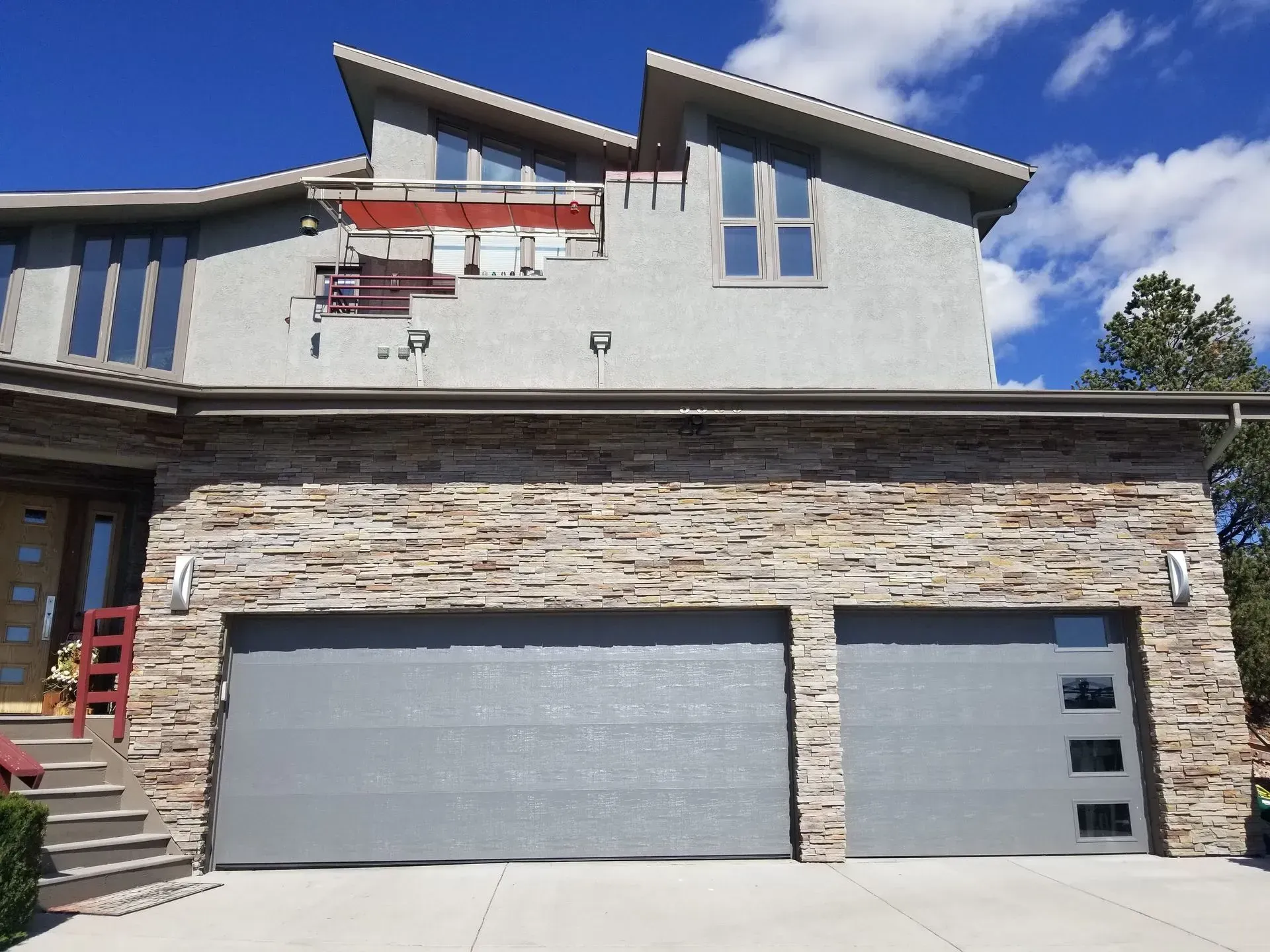 Two-story house with stone facade garage doors, gray, with a section of stucco missing. Blue sky overhead.