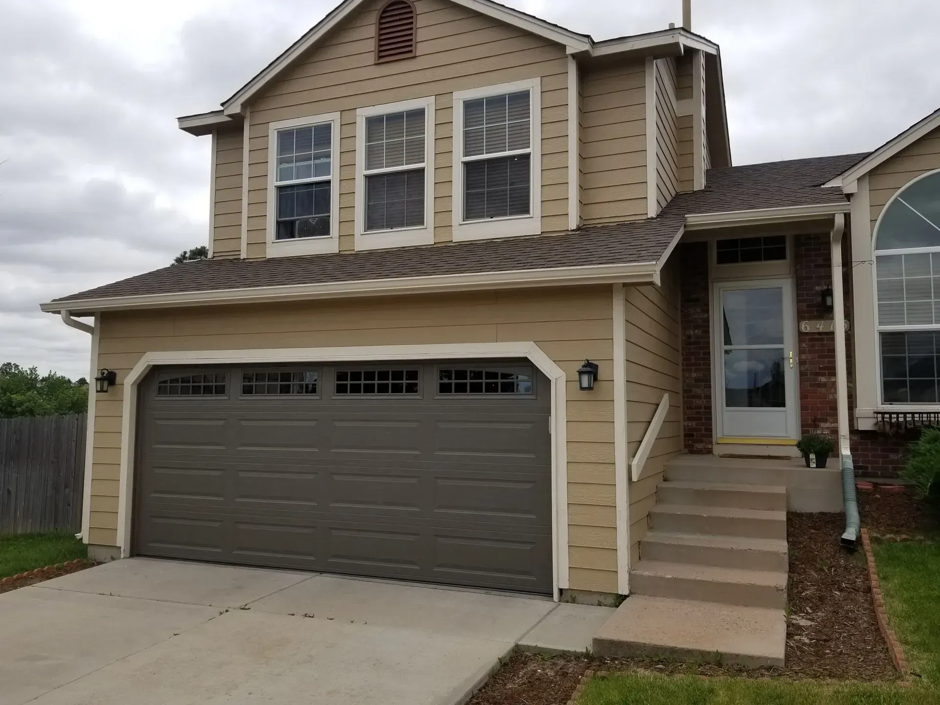 Tan two-story house with a brown garage door and steps leading to the front door under a small roof.