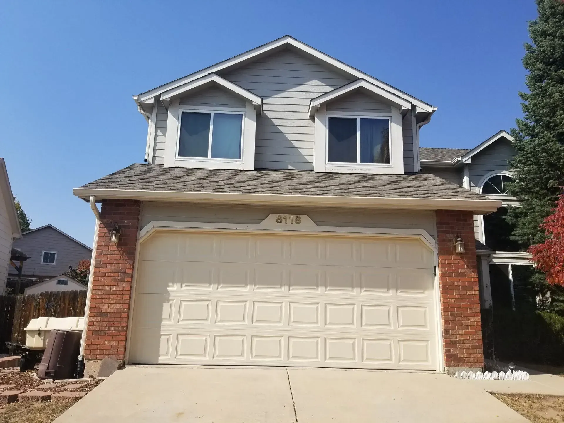 Two-story house with a beige garage door and gray siding. Brick accents frame the garage and front entrance.