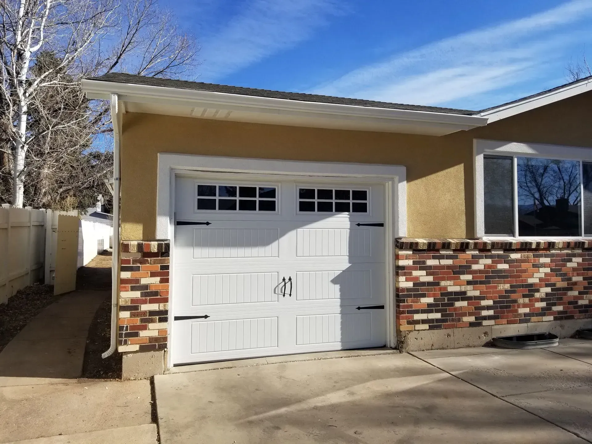 White garage door with decorative hardware on a stucco house with brick accents, sunny day.