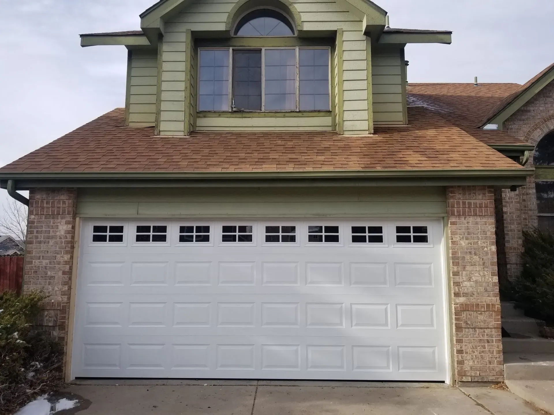 White garage door on a brick and green-trimmed house with a brown roof.
