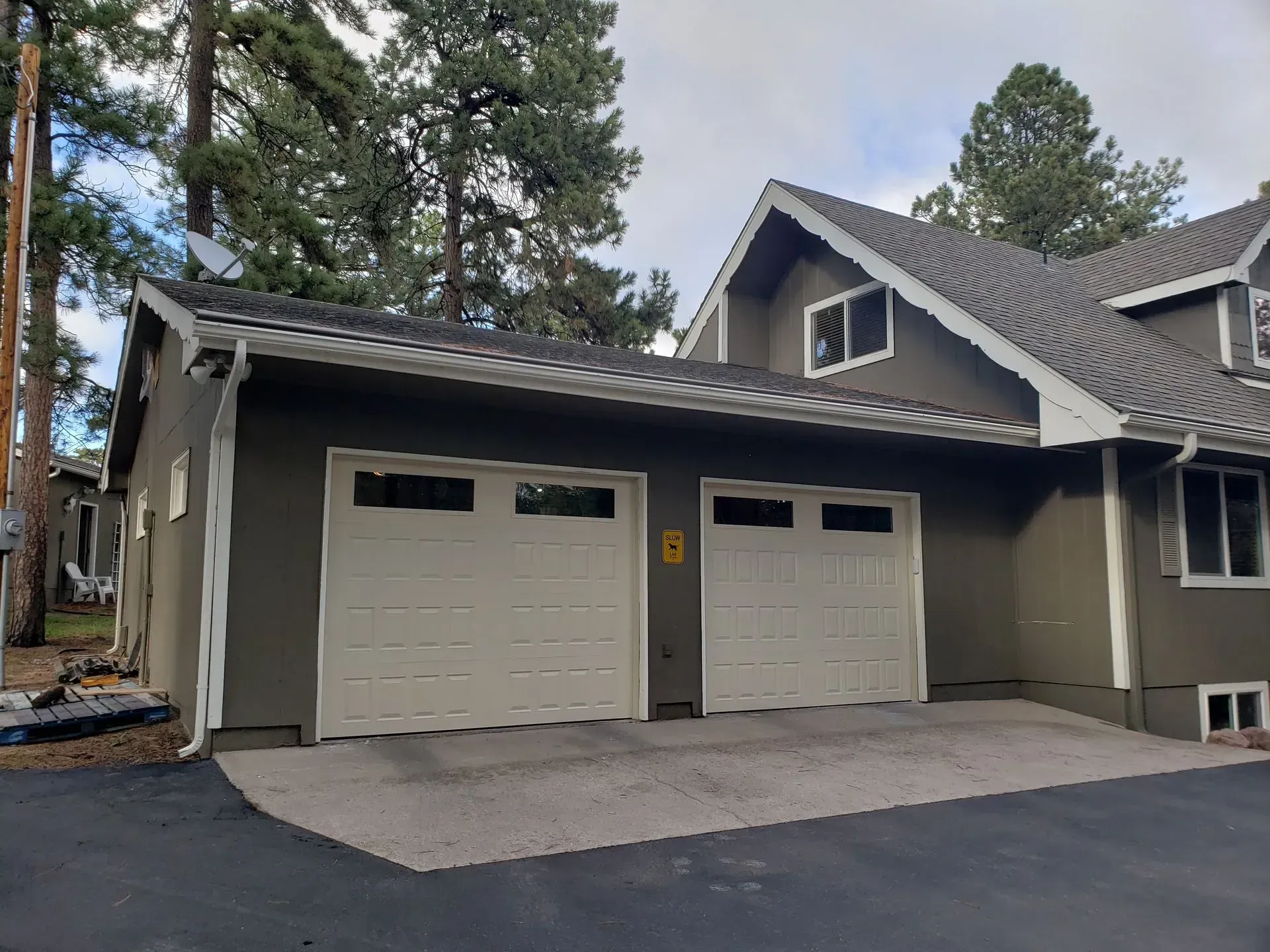 Two-car garage with beige doors on a grey stucco house with asphalt driveway and trees.