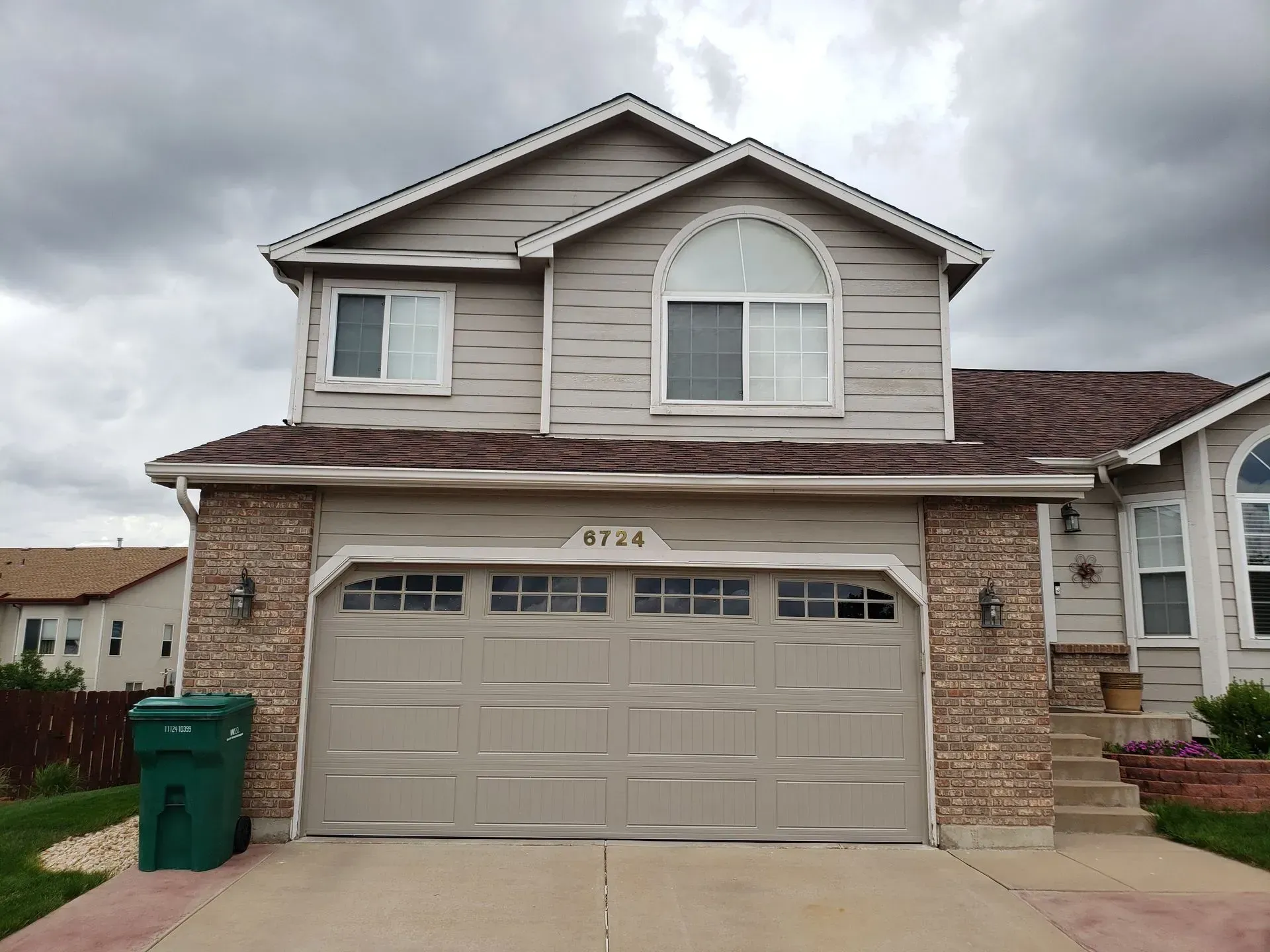 Two-story house with tan siding, brick facade, and a gray garage door; cloudy sky.