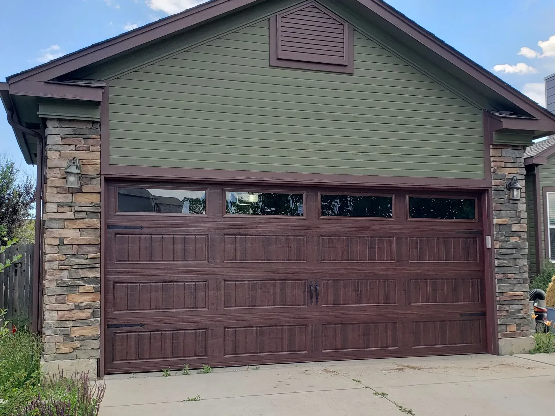 Brown garage door with windows, flanked by stone columns, under green siding.