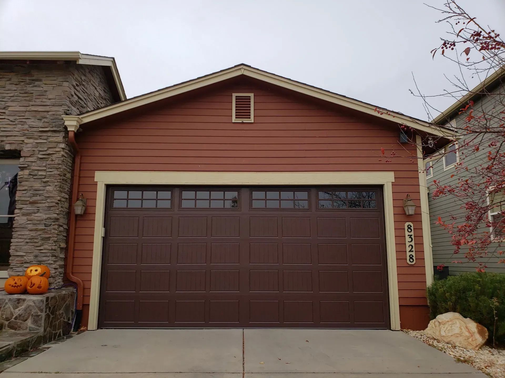 Brown double garage door on a house with red siding and stone accents, flanked by pumpkins and fall decor.