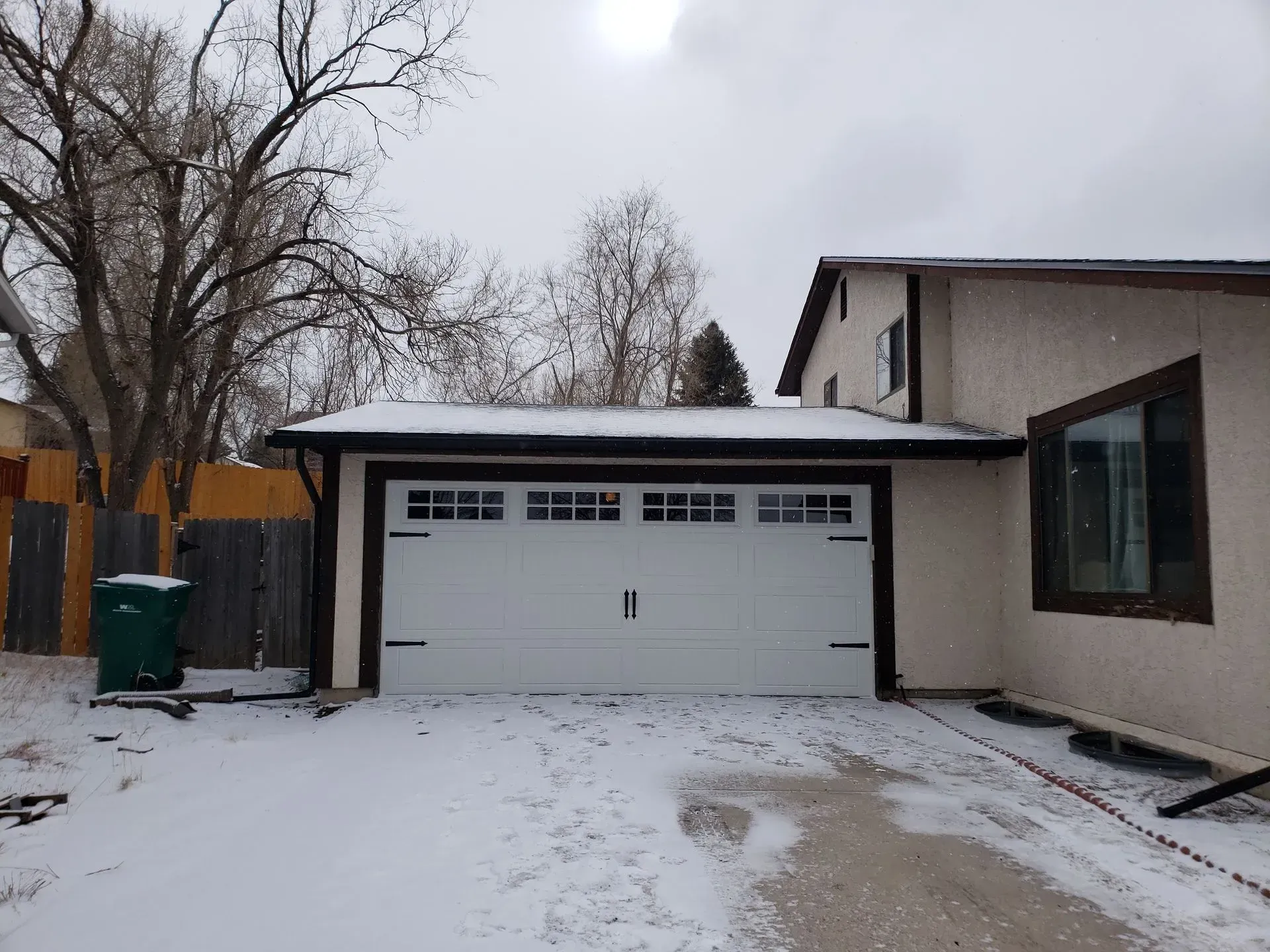 Snowy garage with white door and brown trim, attached to a light-colored house.