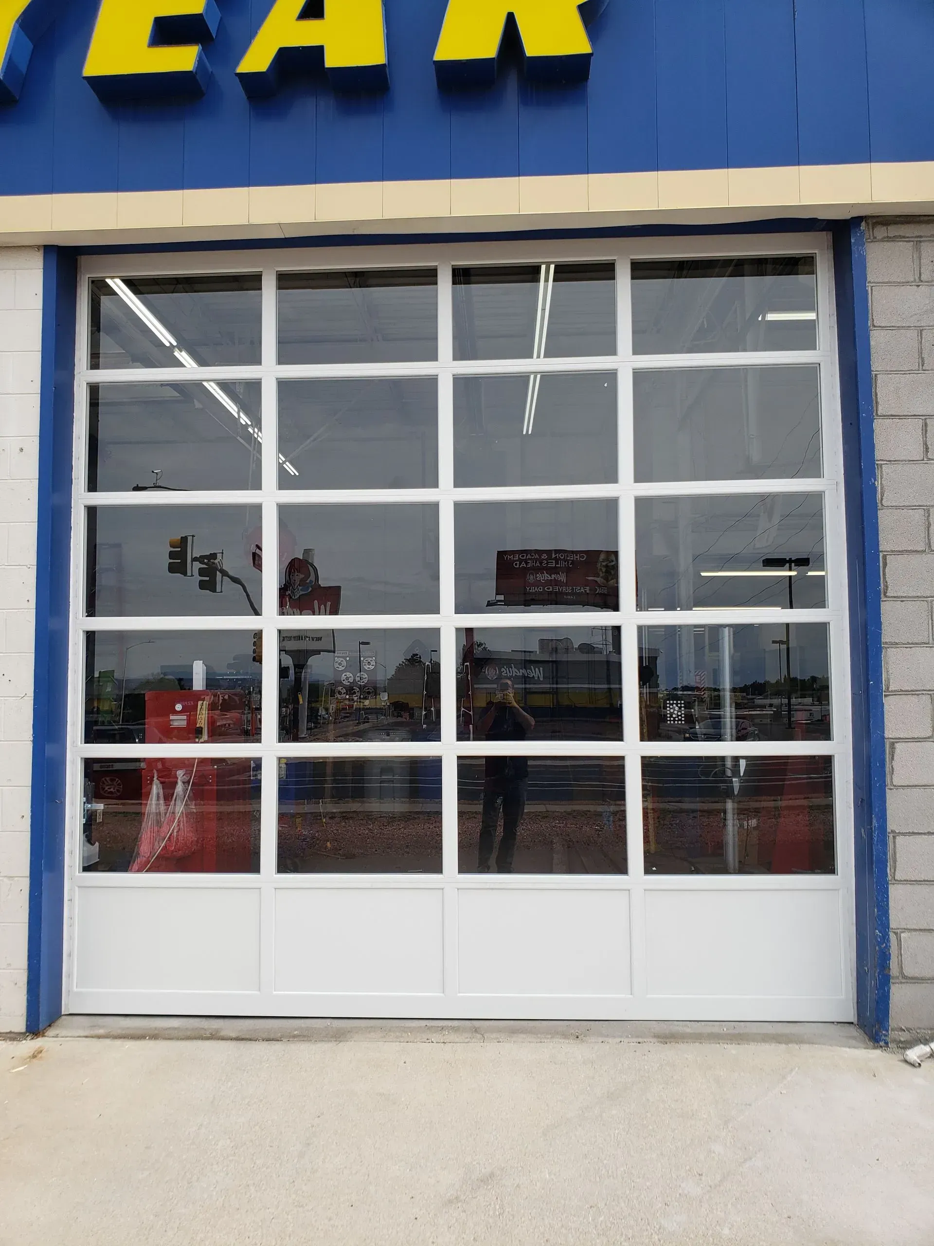 Glass garage door at a Goodyear shop, reflecting street scene. White frame, blue signage above.