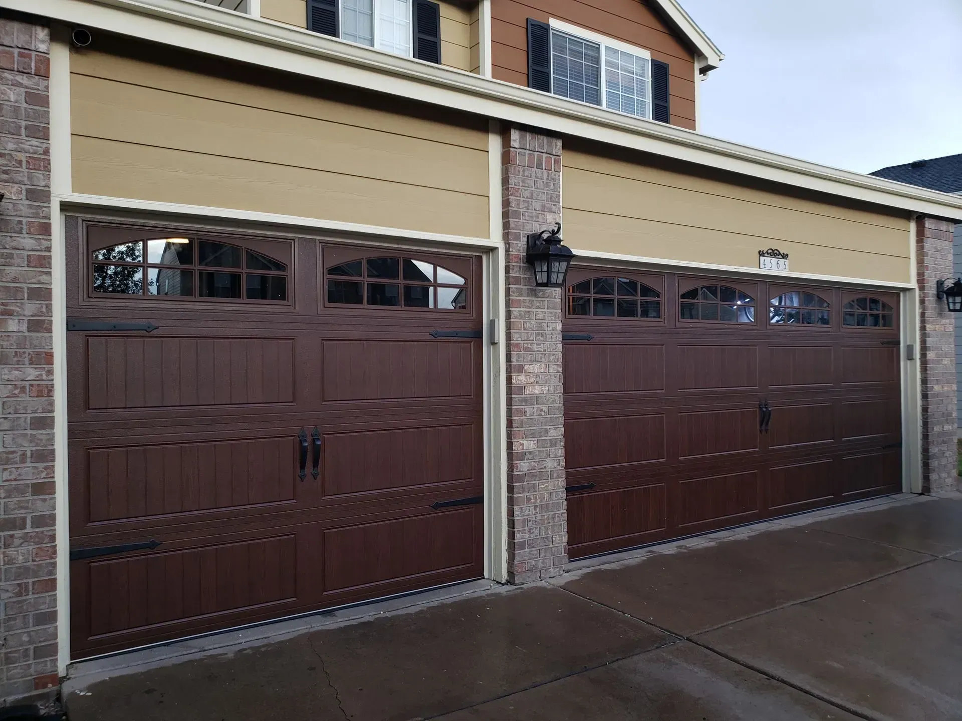 Two brown garage doors on a house with tan siding, brick columns, and a paved driveway.