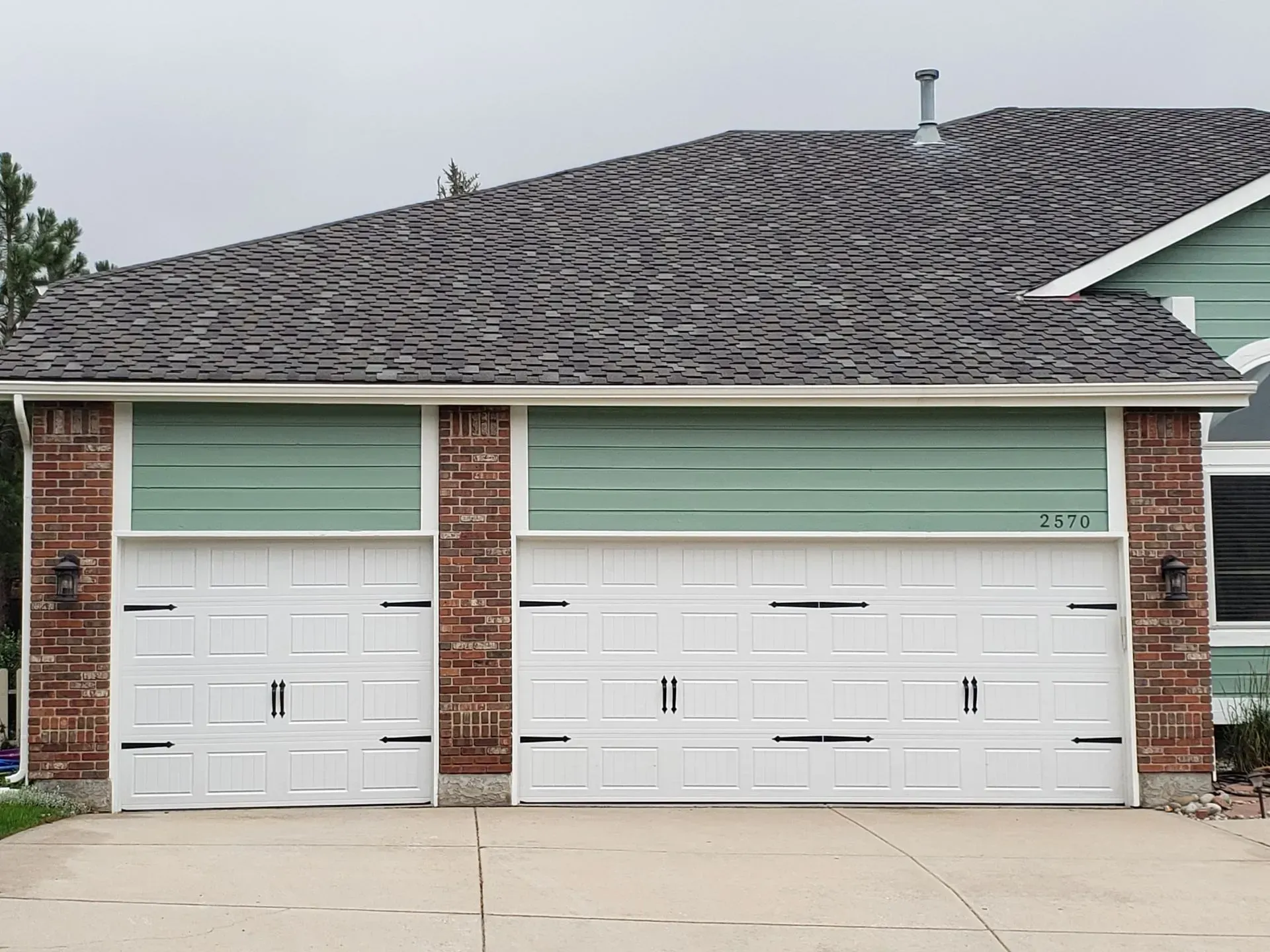 Two white garage doors with green siding above, set in a brick frame, under a gray roof.