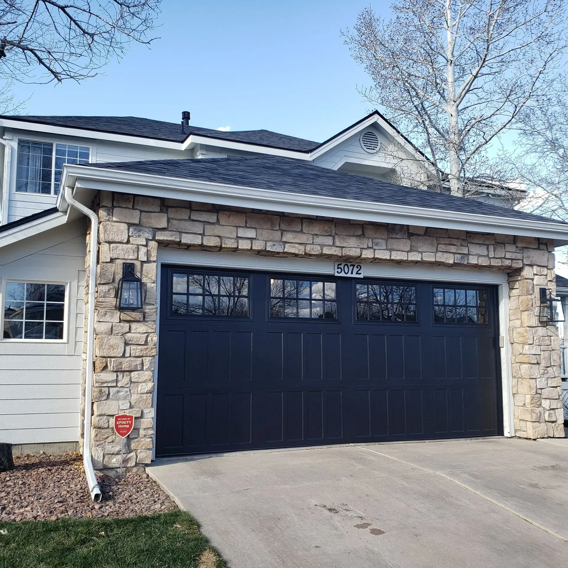 Black garage door with stone facade on a two-story house. White siding, and asphalt roof.