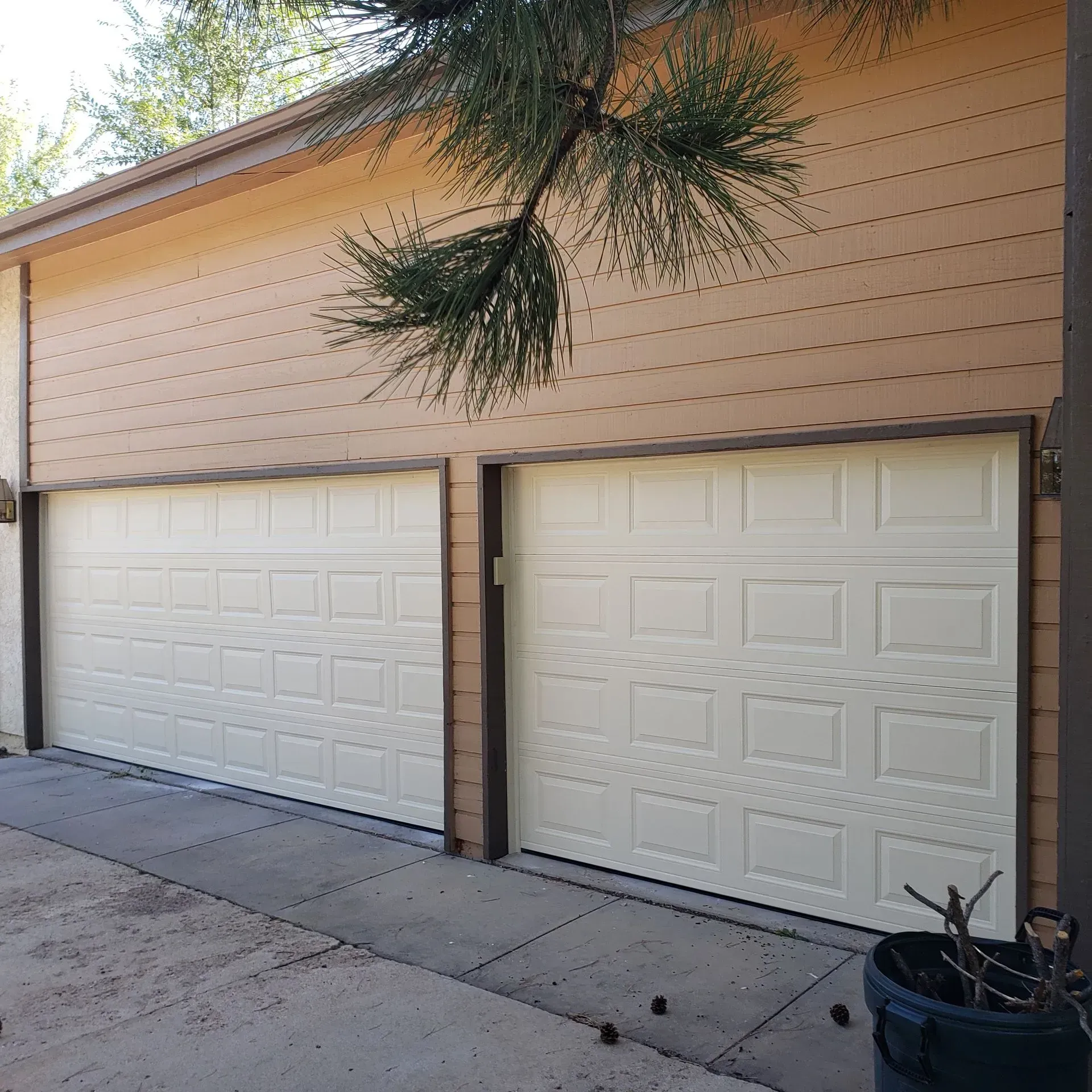 Two cream-colored garage doors with brown trim, under tan siding, tree branches overhead.