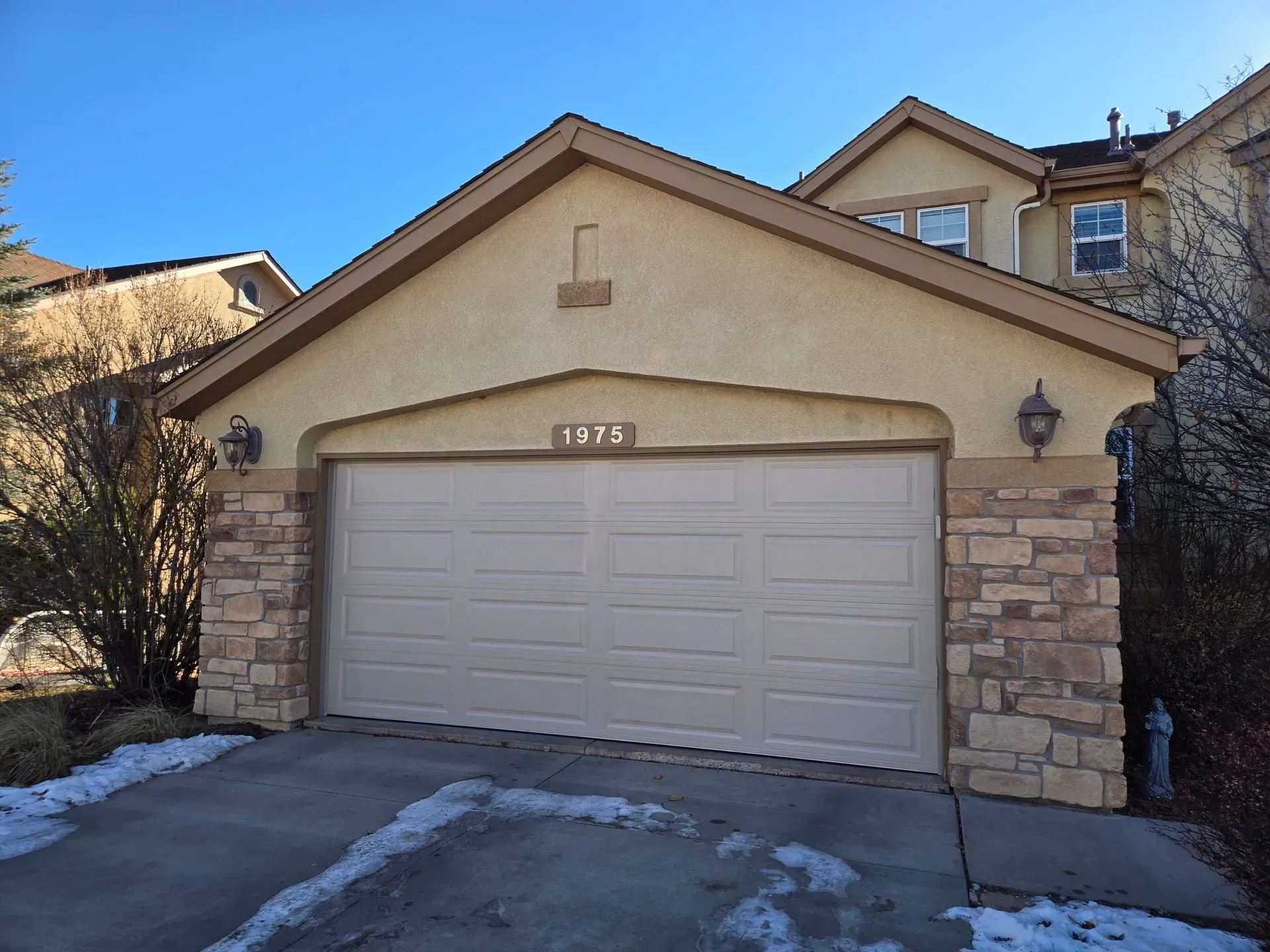 Beige garage door with stone accents, snow on the driveway, under a clear sky.