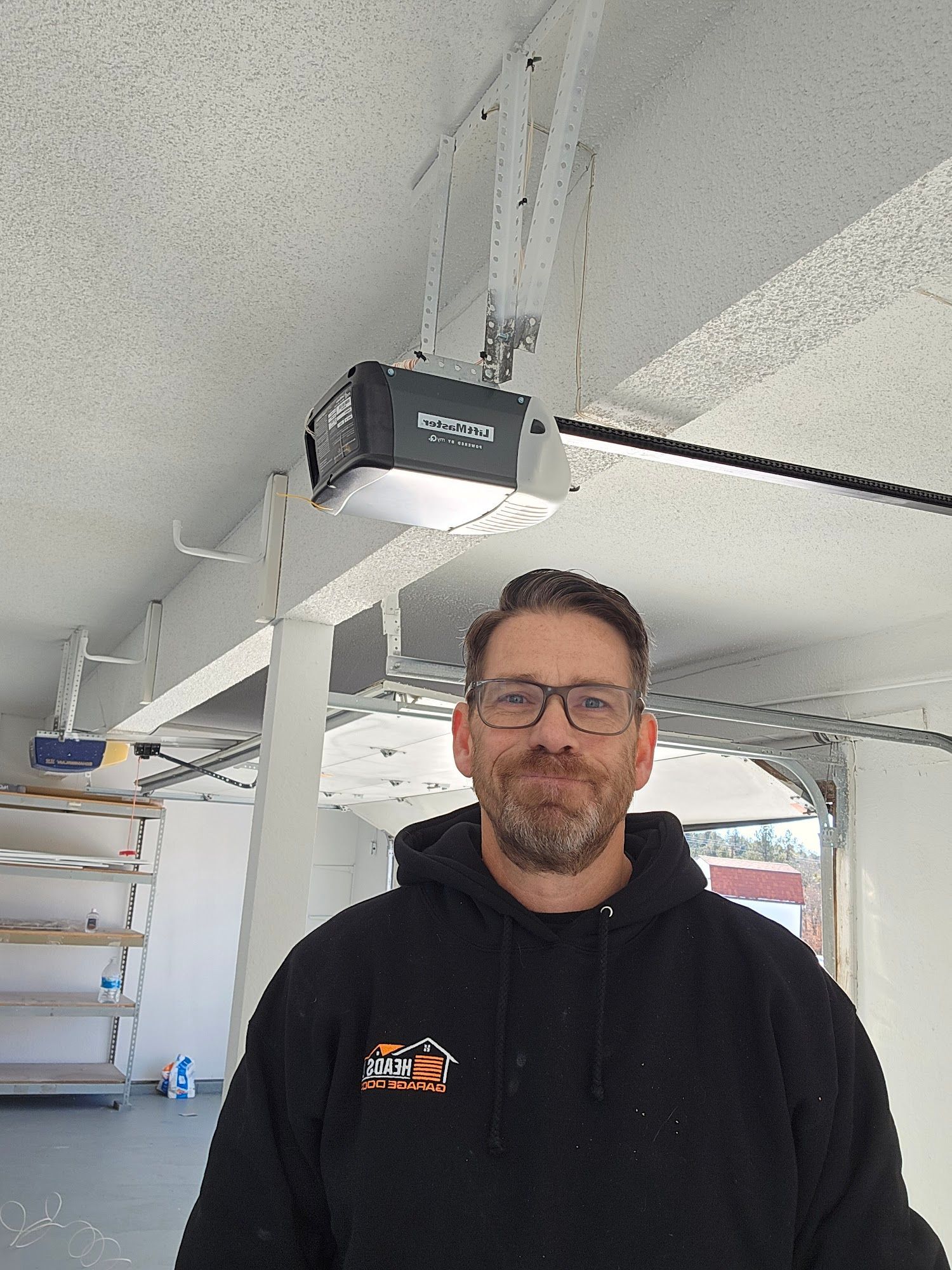 Man in black hoodie stands in a garage; a garage door opener is behind him.