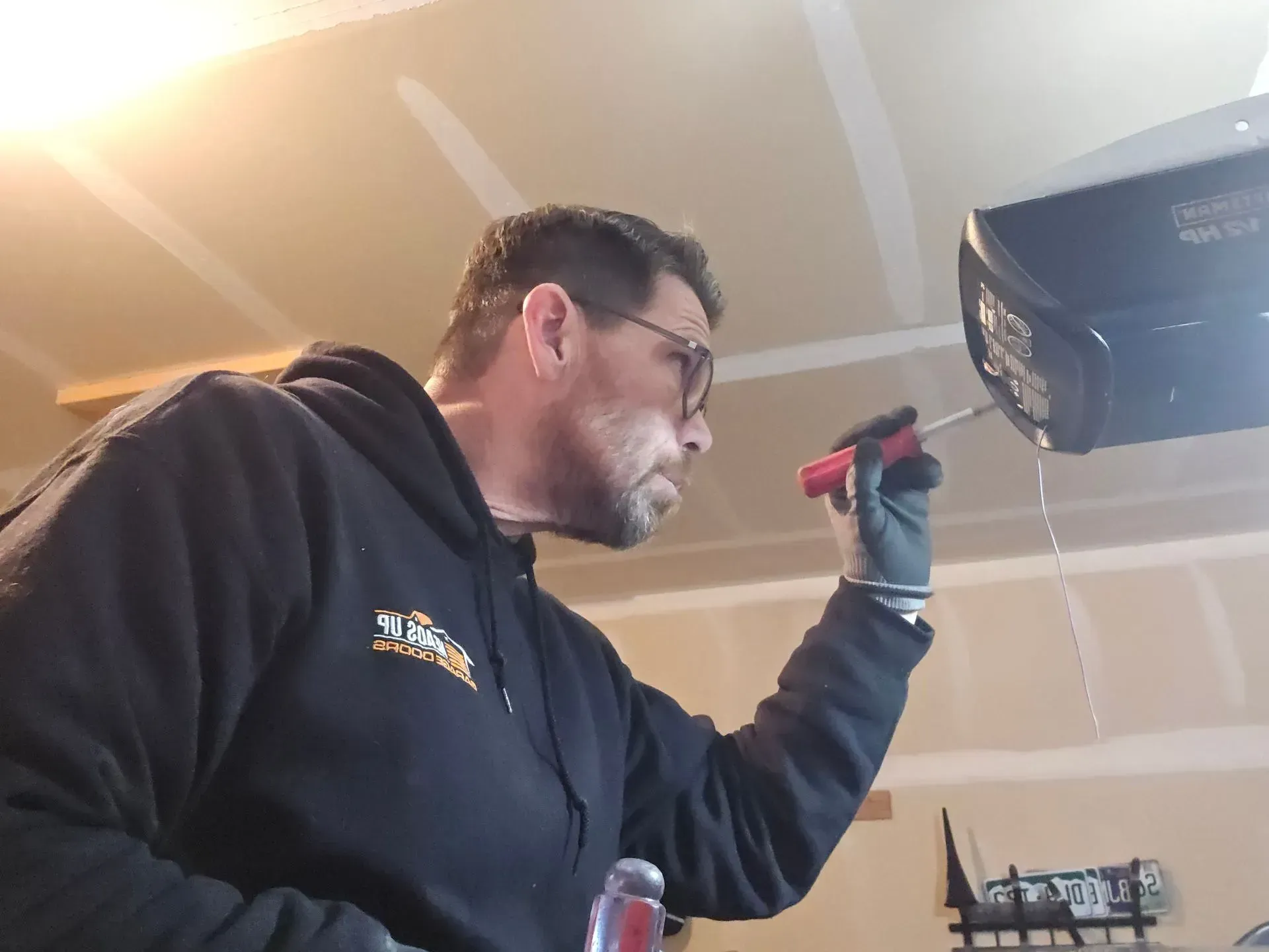 Man in black hoodie, fixing garage door opener with a screwdriver. Inside a garage.